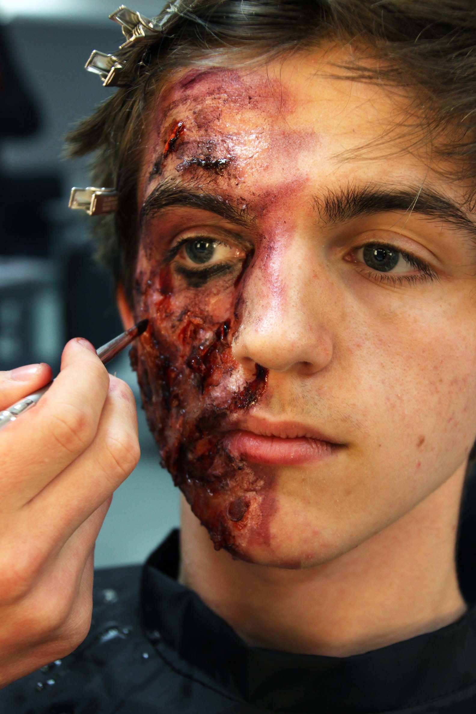 Person having special effects makeup applied to create realistic wound and injury effects on left side of face and forehead.