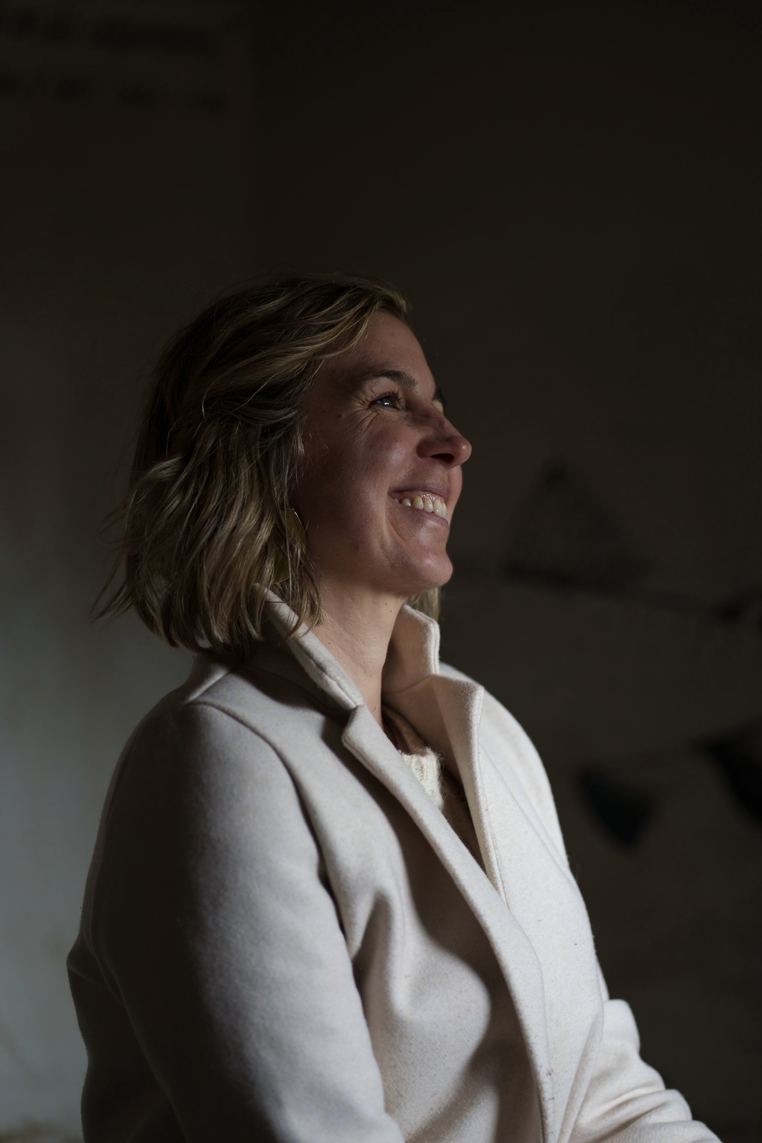 Woman with shoulder-length blonde hair smiling, wearing cream blazer, looking upward in profile against blurred background.