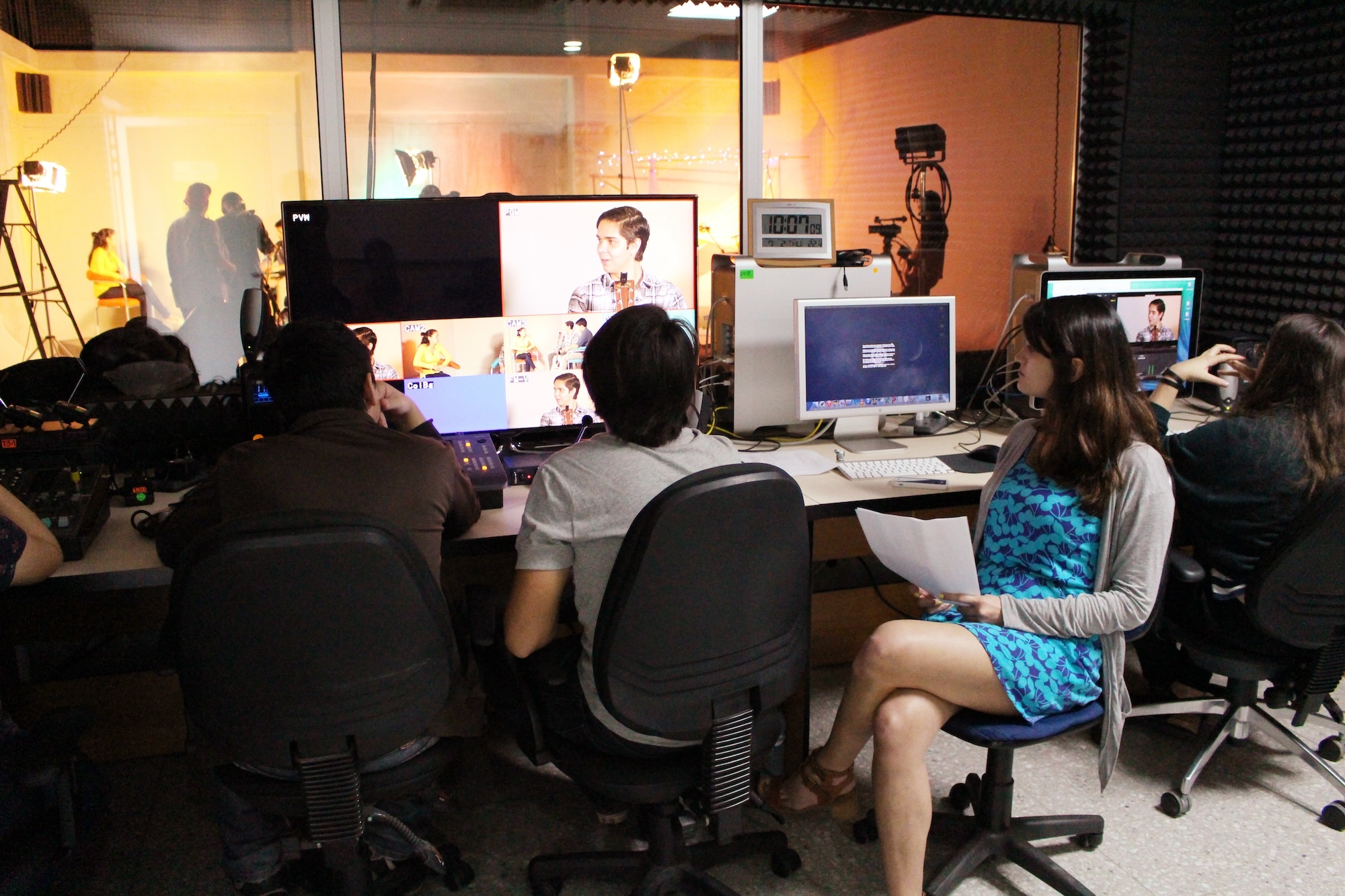 Four students sitting in front of monitors in an editing suite while a shoot takes place in a studio in the background.
