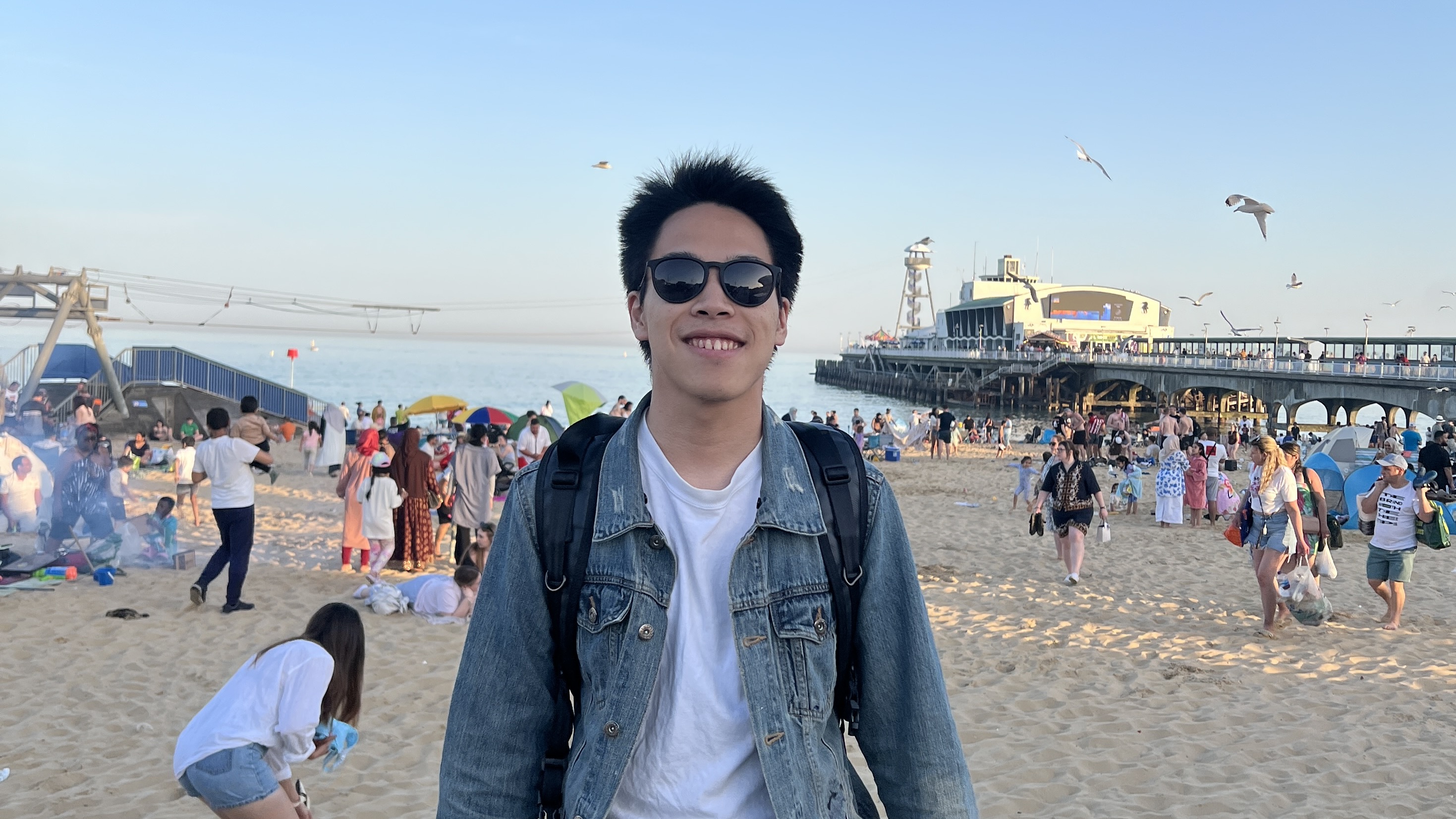 Seiji Suzuki smiling, wearing sunglasses, denim jacket and a bag, on Bournemouth Beach during the evening, seagulls and people in the background.