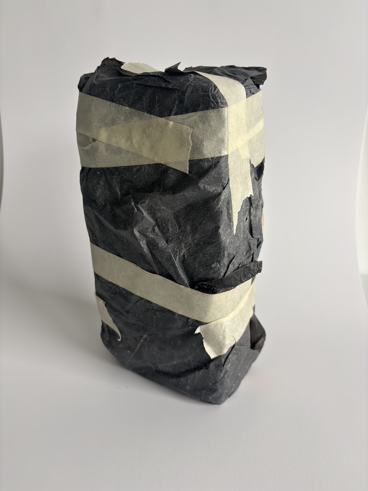 A tall black wrapped cylindrical package secured with beige tape, standing on a white surface against a plain light background.