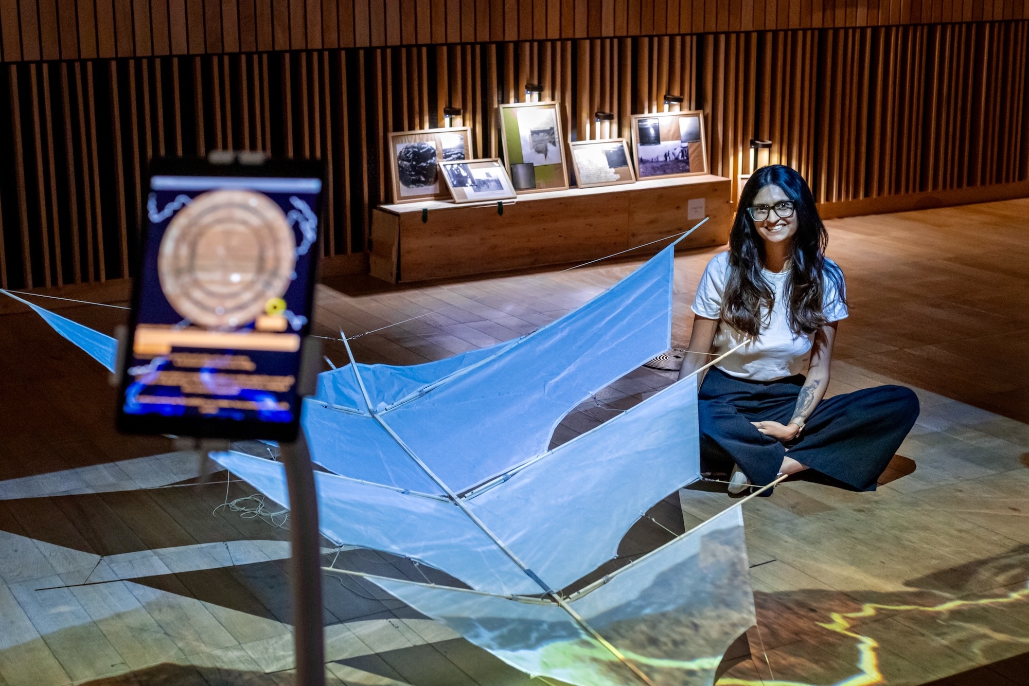 K Saraiya sitting beside large blue geometric glass sculpture in wood-panelled gallery with illuminated display cases.