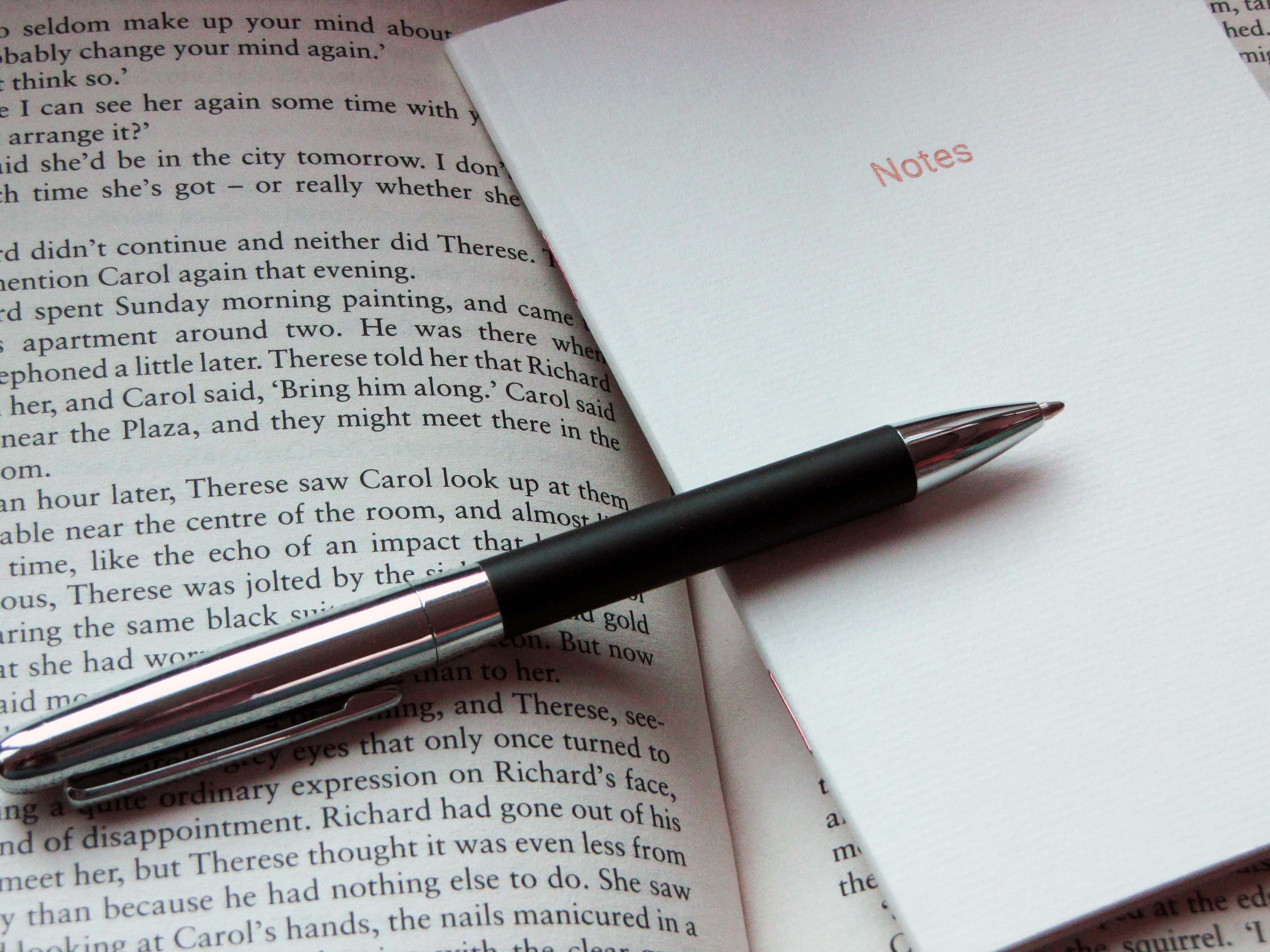 Close-up of an open book, with a note pad and pen lying across the pages.