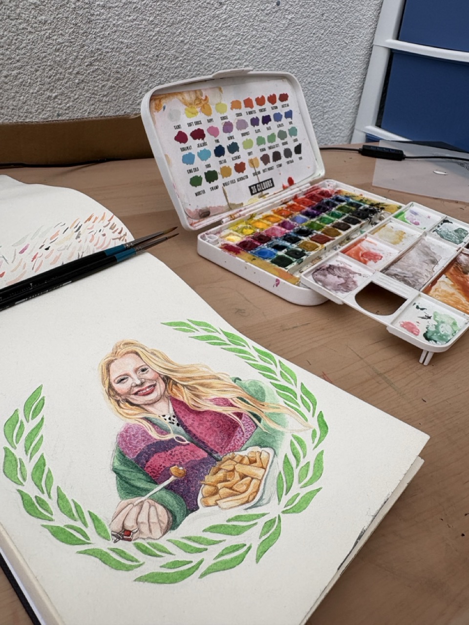 An open sketchbook on a wooden desk shows a coloured illustration of a blond woman holding food and a pumpkin, with an open watercolour paint set beside it.