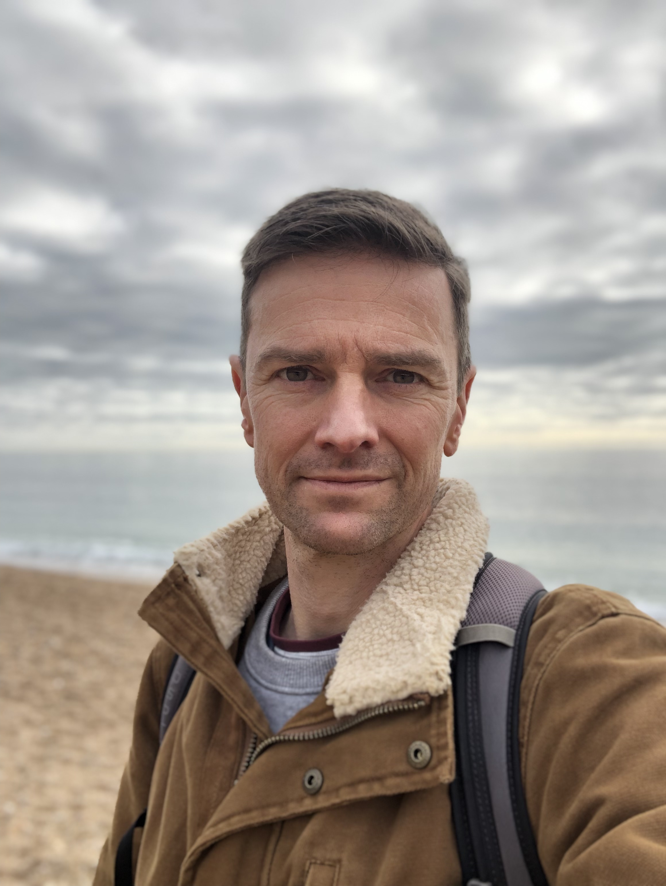 Man in brown jacket with fleece collar taking selfie on beach with cloudy sky and sea in background.