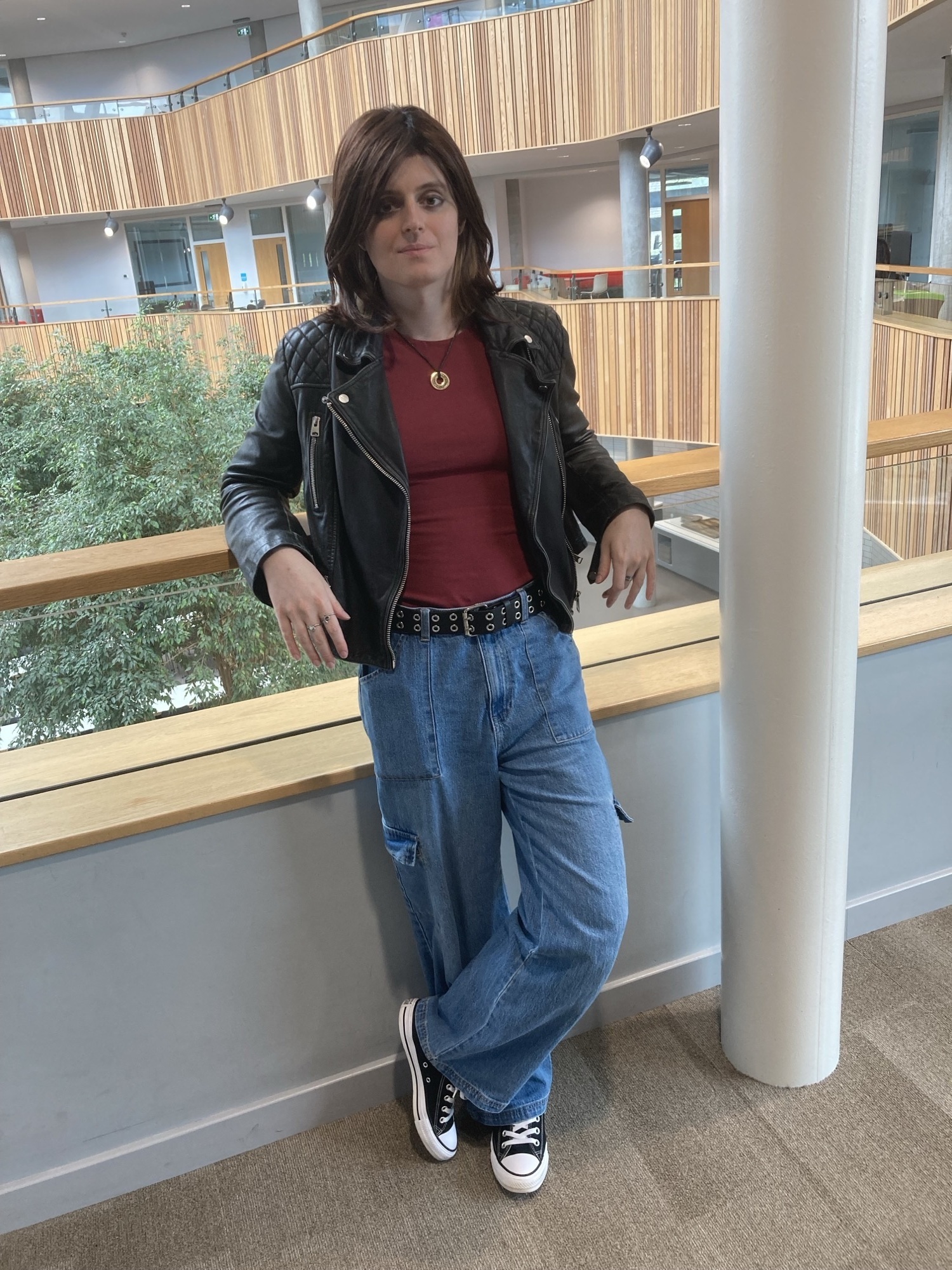 Woman in black leather jacket, red top, blue jeans and black trainers leaning against wooden railing in modern building atrium.