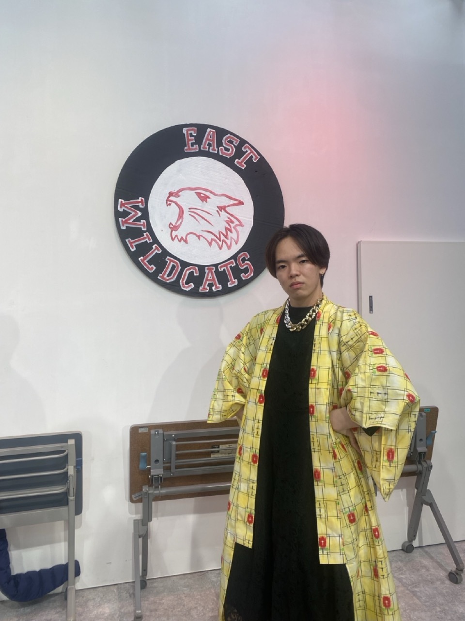 Mitsuki Kawase stood in a flowing yellow and black garment with chains, 'East Wildcats' drawn logo hung on the wall in the background.