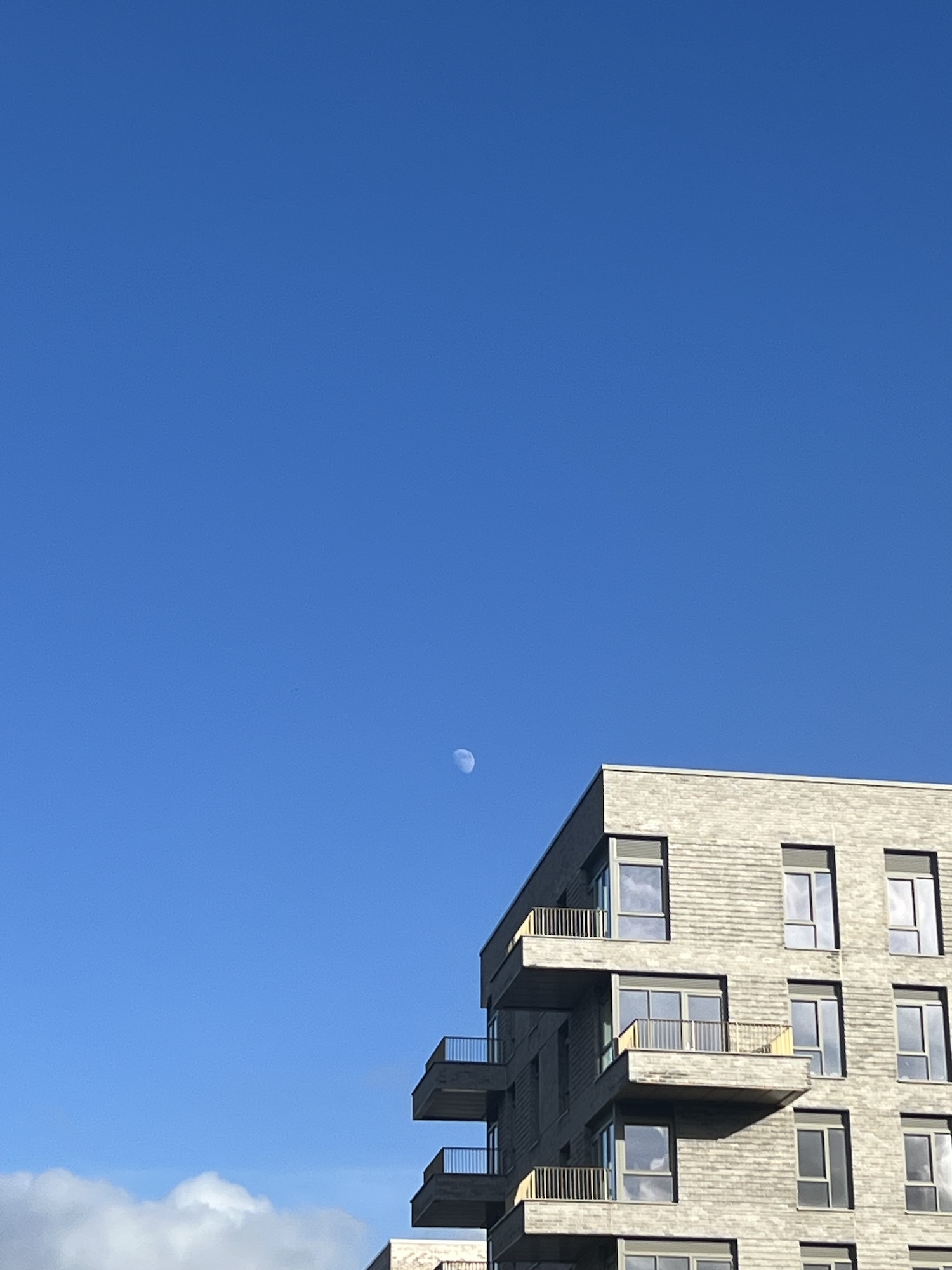 Modern brick apartment building with protruding balconies against bright blue sky with visible moon and white clouds.
