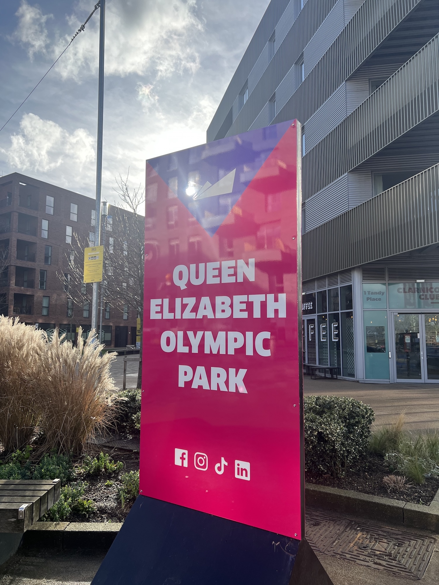 Large pink and purple gradient sign reading "Queen Elizabeth Olympic Park" with social media icons, positioned between modern buildings.