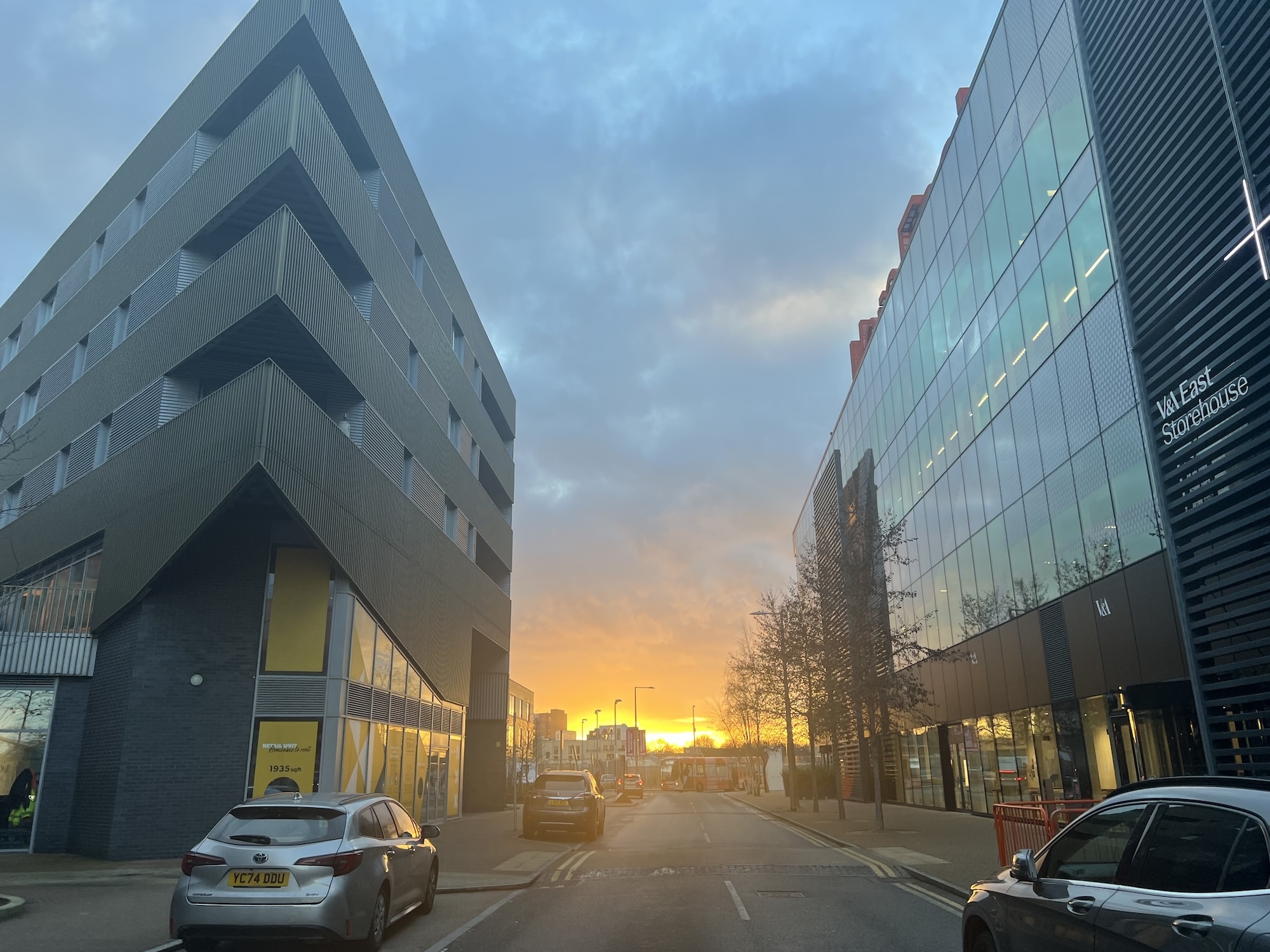 Modern concrete and glass buildings line urban street at sunset with orange sky, parked cars, and bare trees visible.