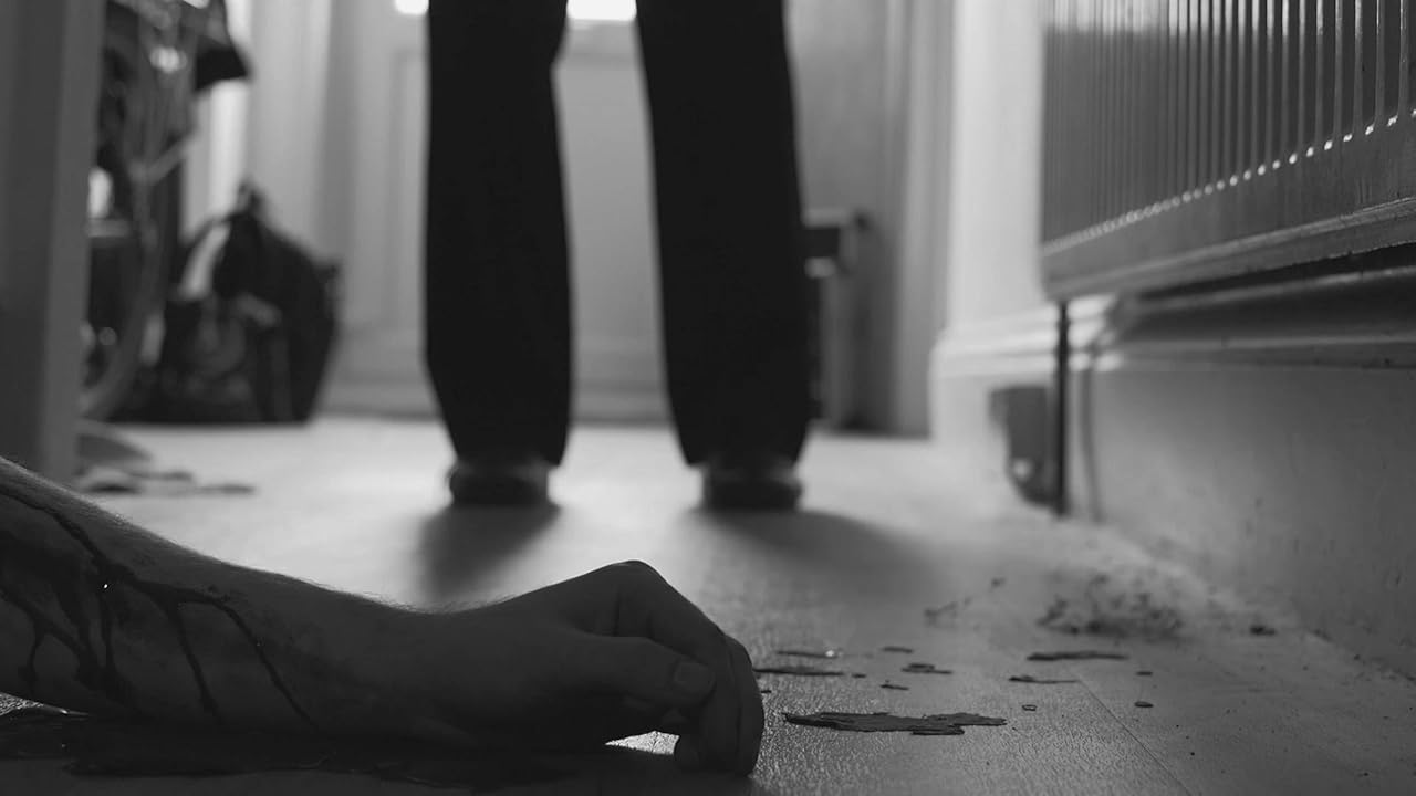 A person lies on the floor in a hallway, with another person standing in the background near a radiator.