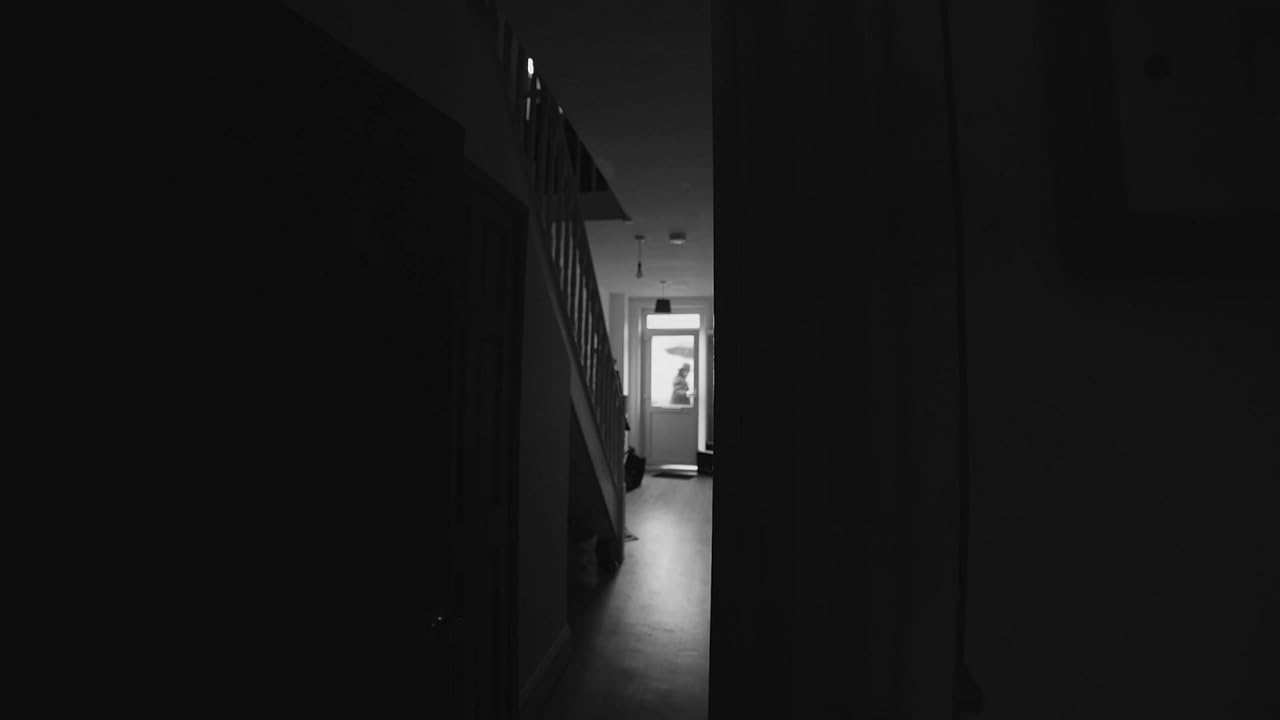A narrow, dimly lit corridor in black and white, with a staircase on the left and a bright doorway at the far end.