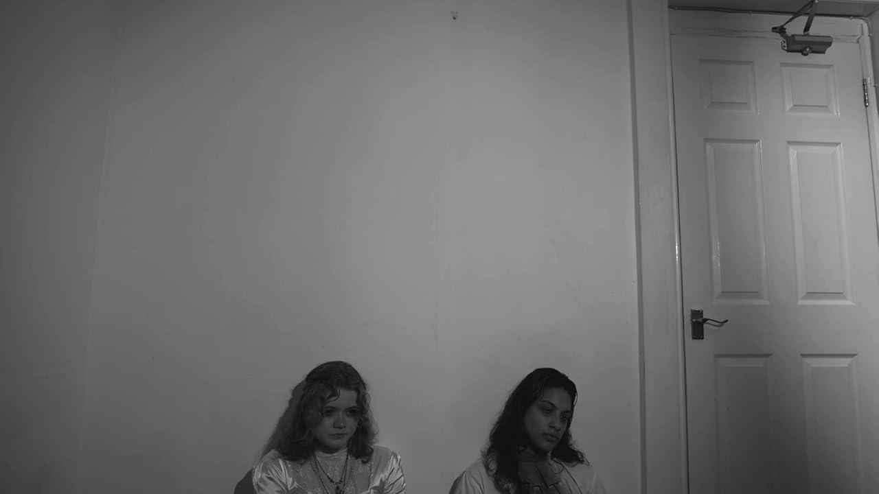 Two people sit on the floor against a plain wall, with a closed door on the right and a bare room around them.