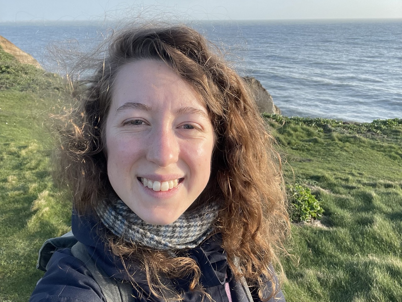 Selfie of Martha Grubb, smiling with wavy brown hair, stood on a grassy cliff with the sea in the background.