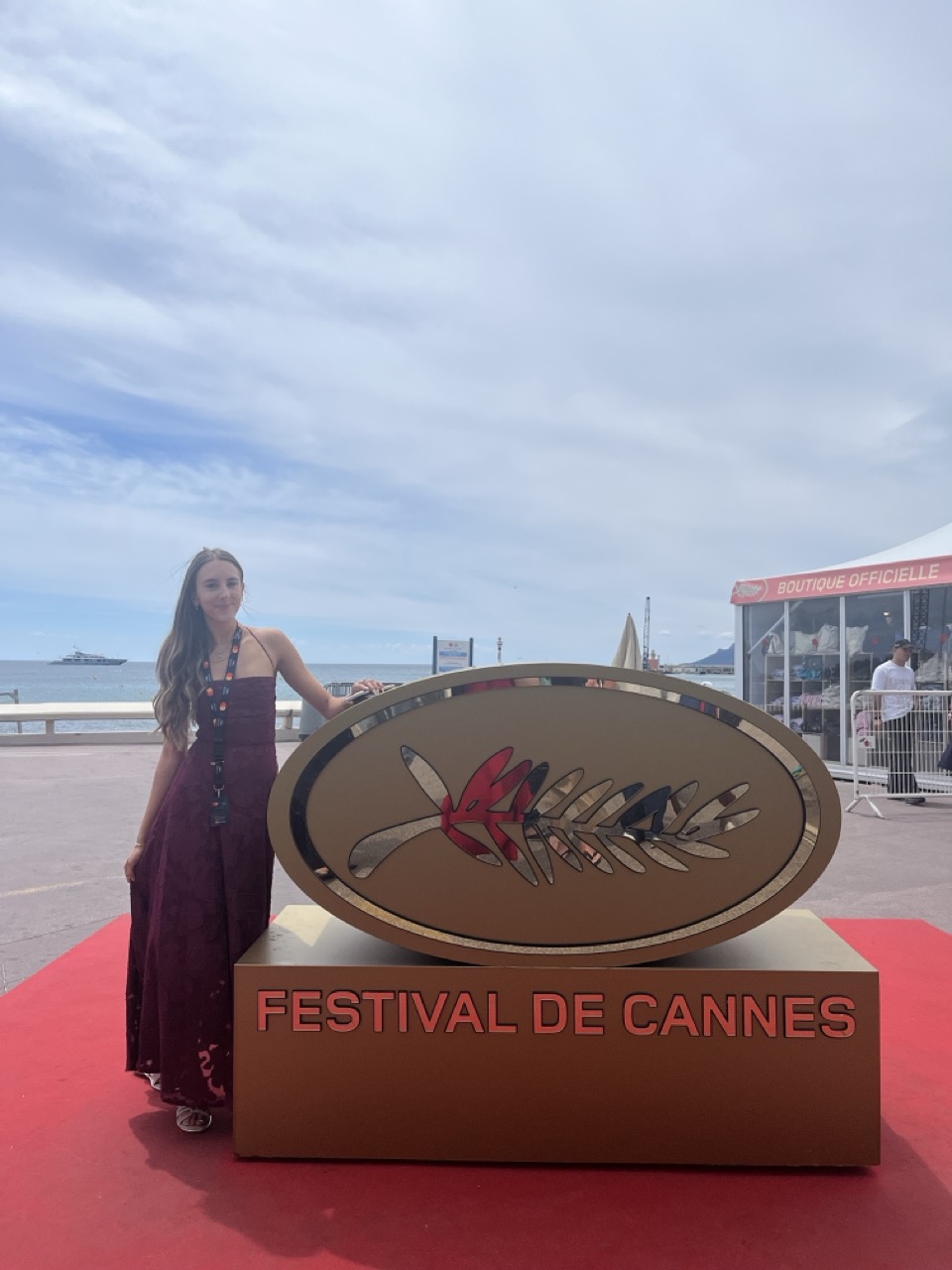 A woman in a burgundy gown stands beside a large Cannes festival emblem on a red carpet, with the sea and cloudy sky in the background.
