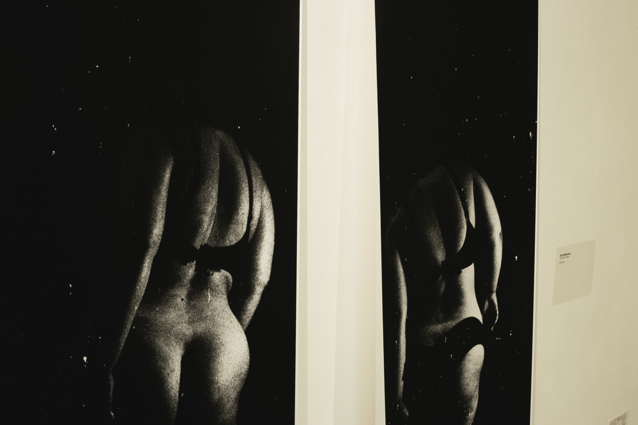Two nude figures visible through glass panels separated by white frames, dimly lit against dark backgrounds.