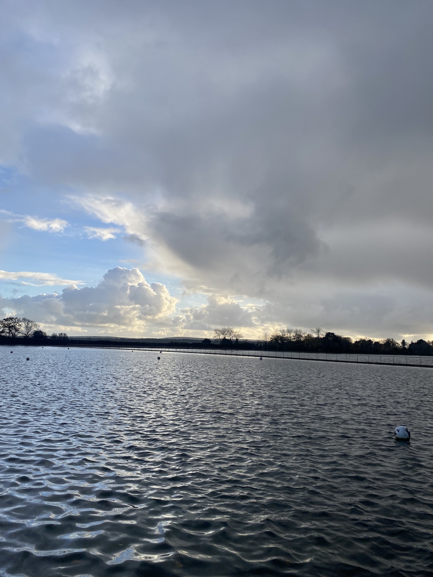 Lake with rippled water surface under dramatic cloudy sky with grey and blue tones. Dark tree line silhouetted on horizon.