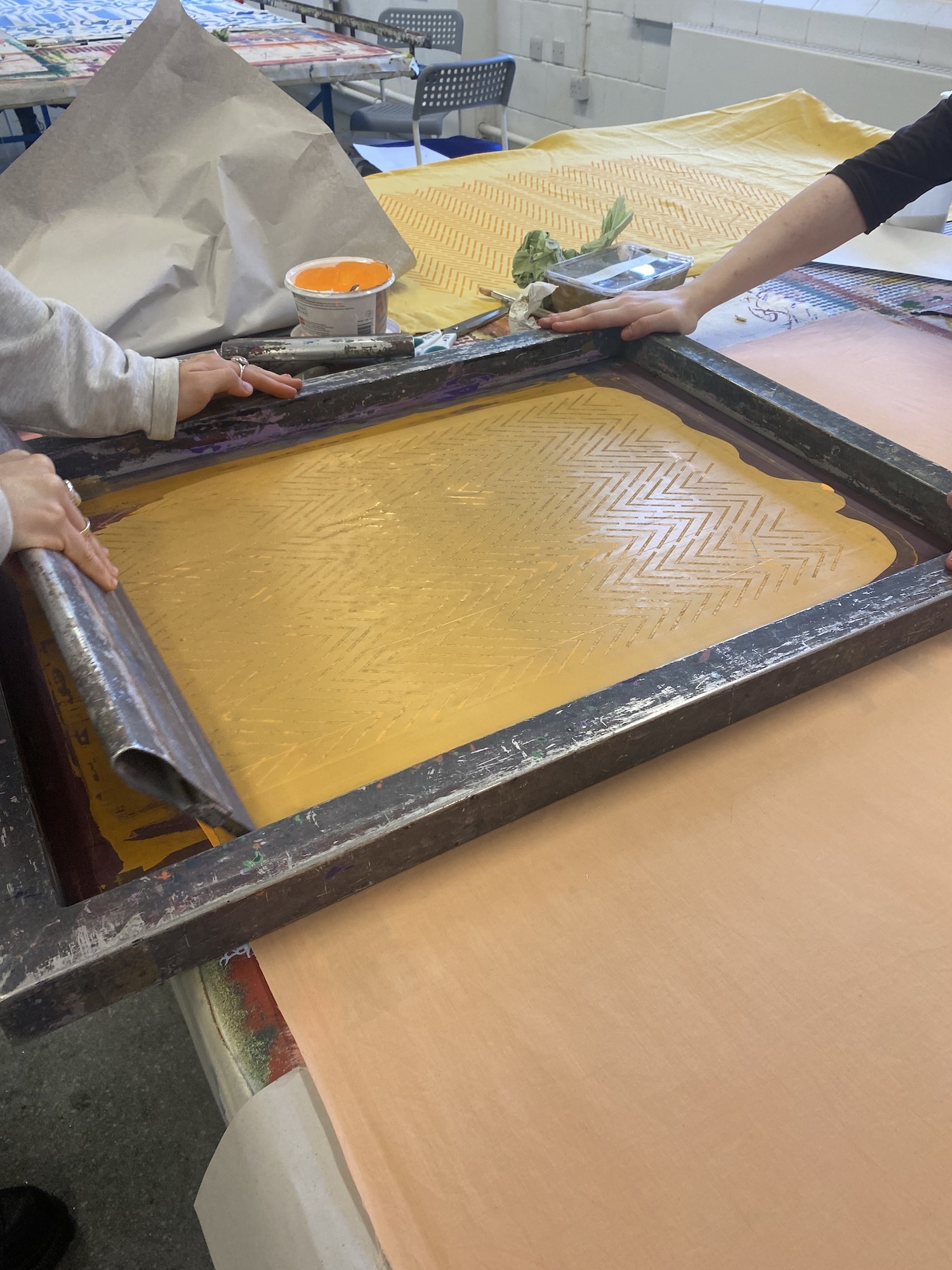 Person screen printing yellow ink onto material using squeegee on printing frame in workshop setting with orange paint pot visible.