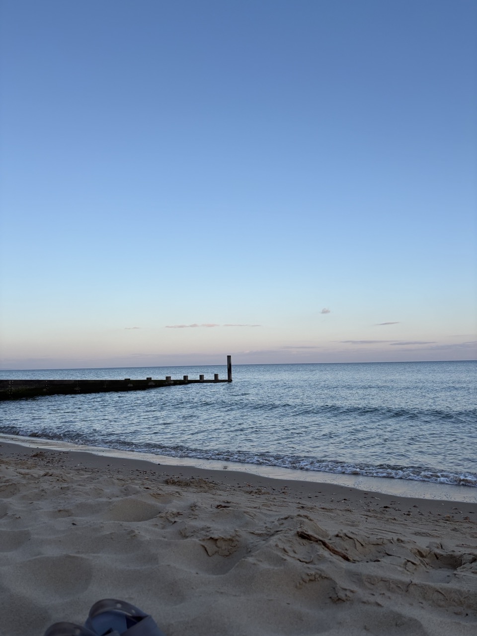 A sandy beach beside calm sea water at dusk, with a long groyne extending into the water under a clear blue sky.