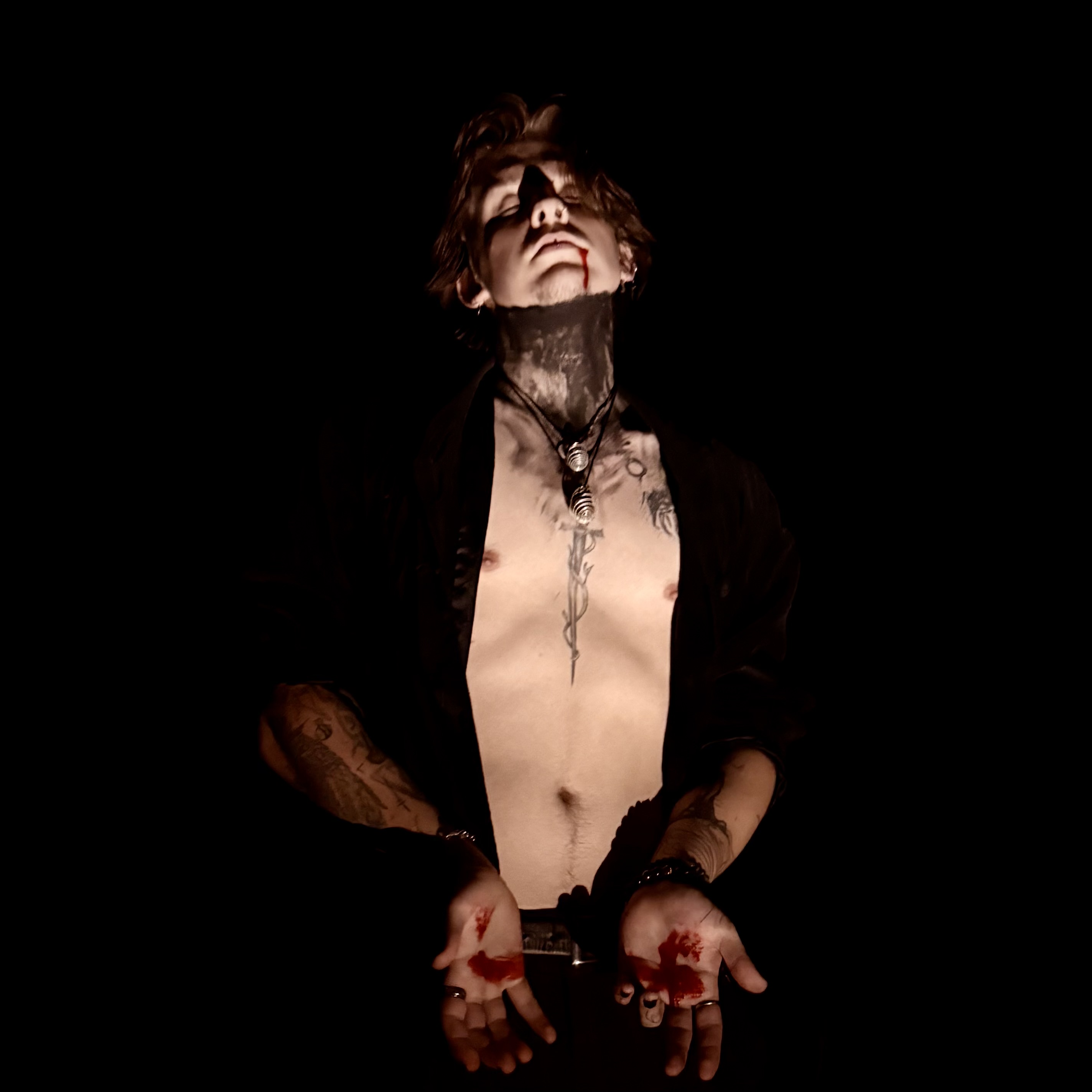 A tattooed person with blood on their hands stands against a black background, wearing an open dark jacket.