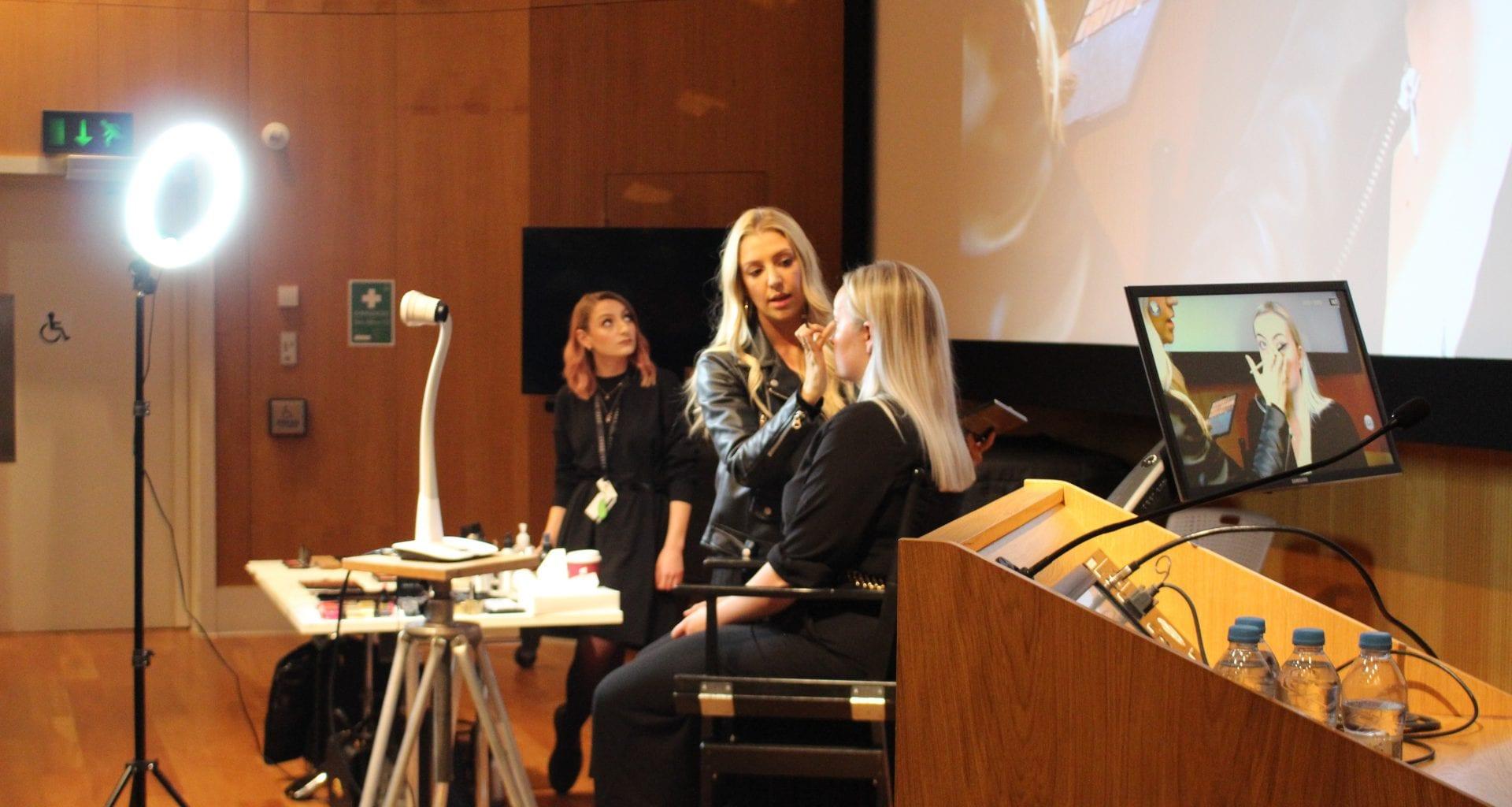 A make-up artist applies make-up to a person sitting in a chair in a lecture theatre.