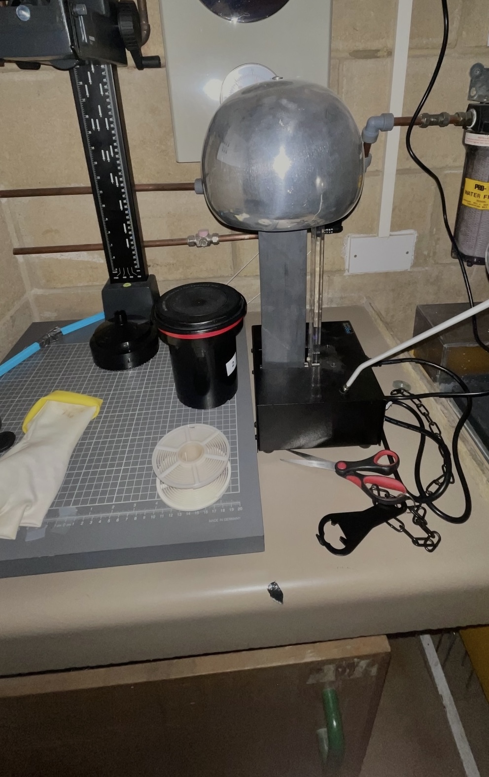 Laboratory setup with black machine, clear glass dome apparatus on stand, petri dish, and various cables on white bench.