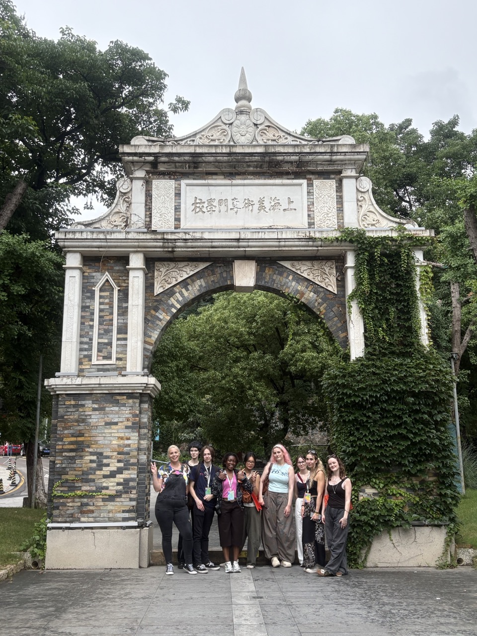 A group of women stands beneath an ornate stone archway with Chinese characters, surrounded by trees and ivy-covered walls.