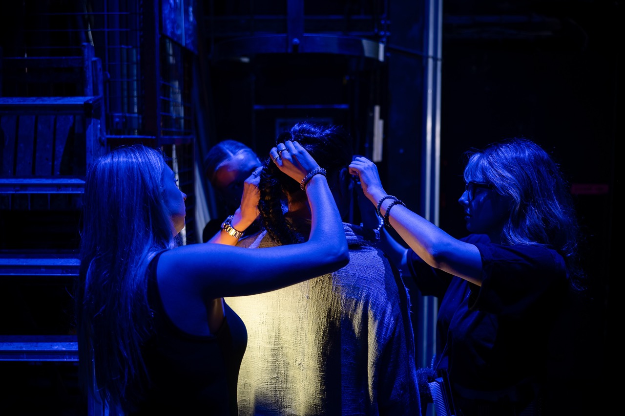 Two women adjusting a person’s hair under blue stage lighting, with a dark backstage area and metal steps in the background.