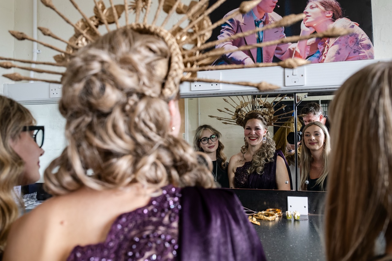 A woman in a purple sequined dress and ornate headdress is seen from behind, facing a mirror with three women reflected in a dressing room.