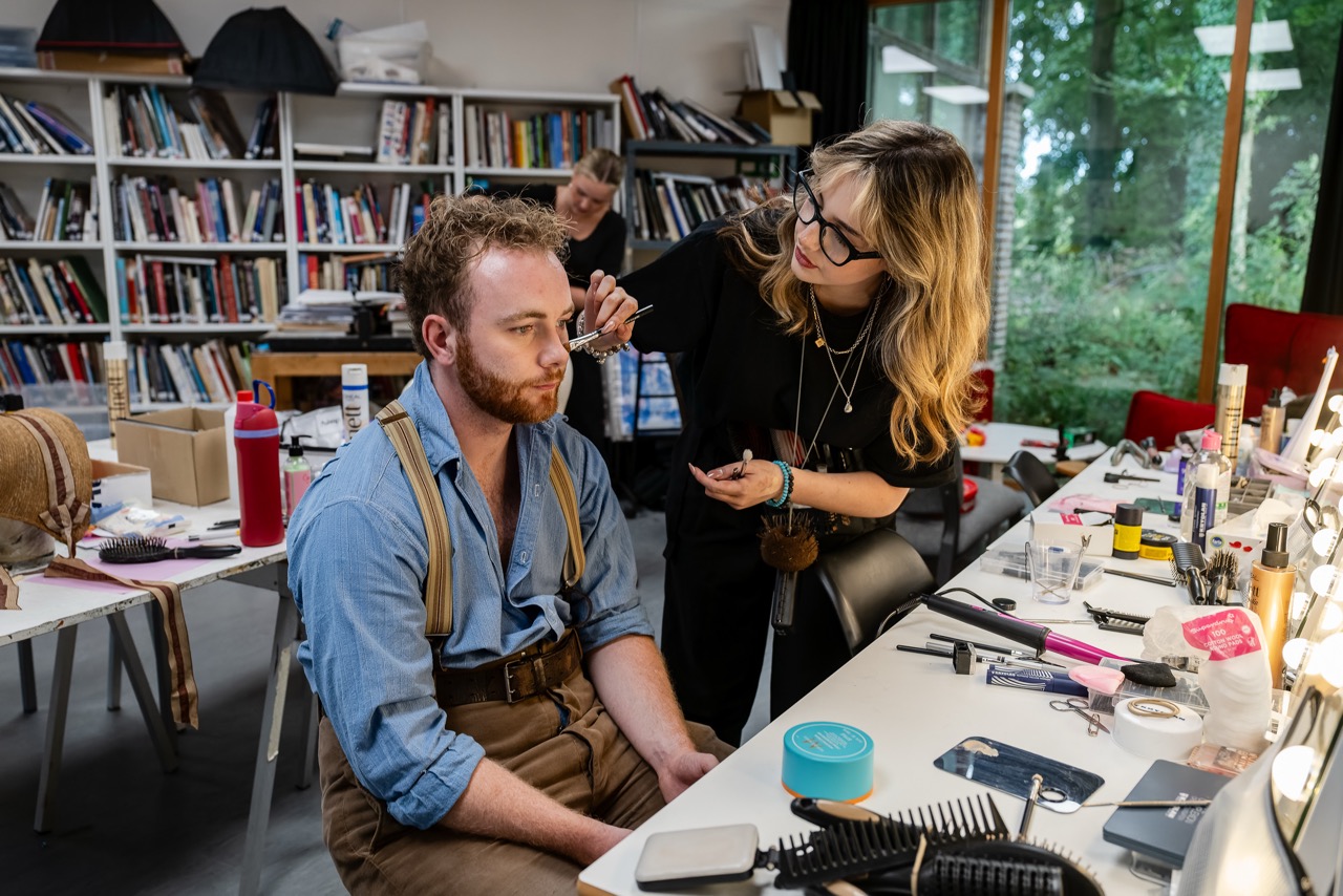A make-up artist applies products to a seated man’s face at a cluttered workstation, with bookshelves and large windows in the background.