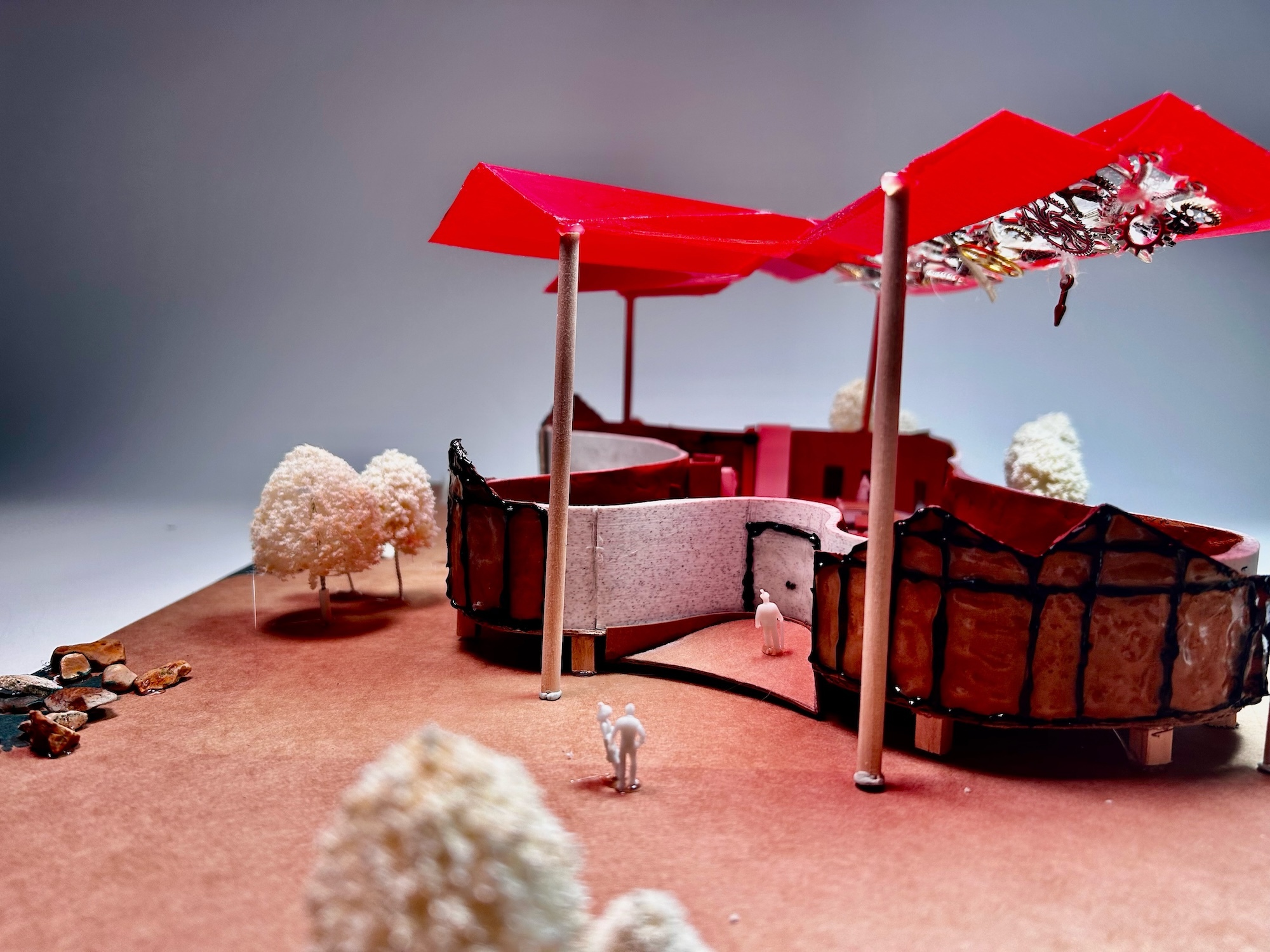 Red fabric canopies on metal poles above pink sandy surface with white coral-like objects and small figurines against grey backdrop.