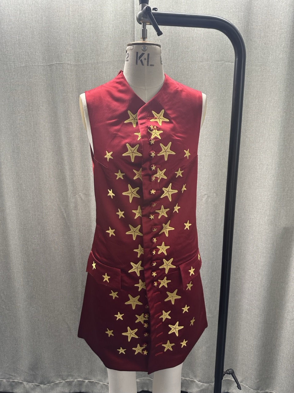 A red sleeveless dress with gold star patterns on a mannequin, displayed beside a black clothing stand against a grey curtain.
