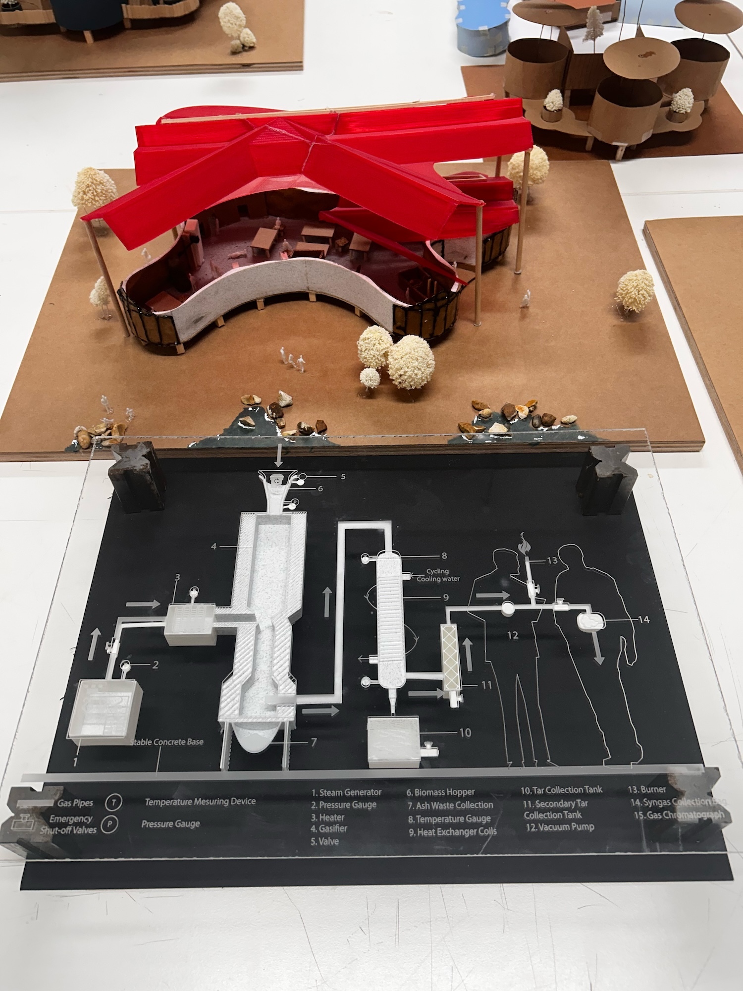 Architectural model with red curved roof structure on brown base, positioned above technical drawing with white building schematics on black background.