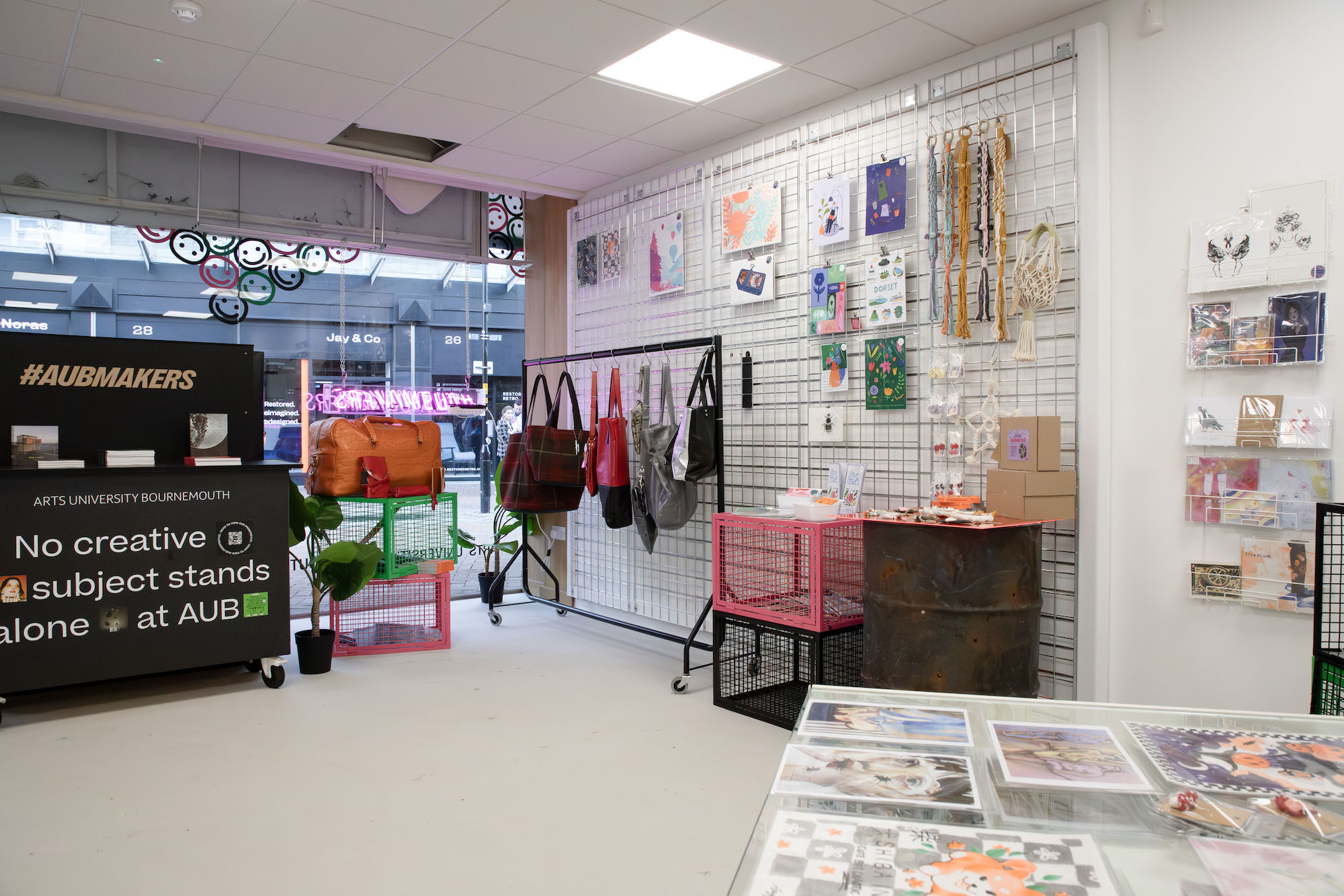 Interior of the AUBMakers pop-up shop with a large metal rack hanging on one wall, with several products on display, and a clothes rail, mesh crates and a barrel in front of it, also with products on display on them.