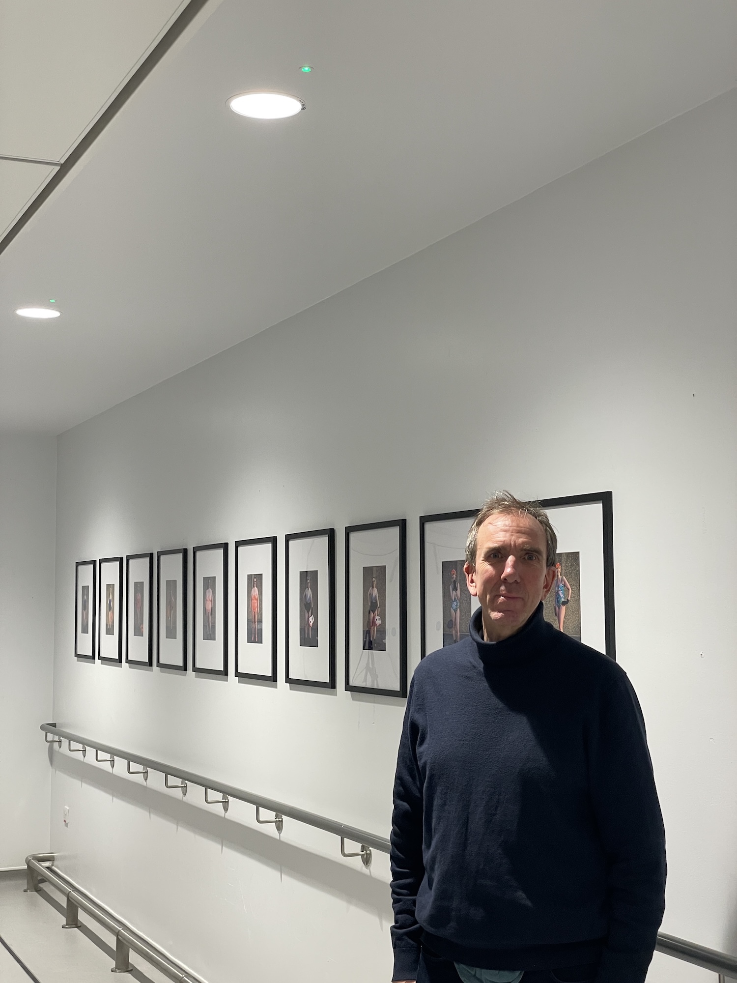D Bird standing in white gallery beside row of black-framed artworks mounted on wall with metal handrail below.