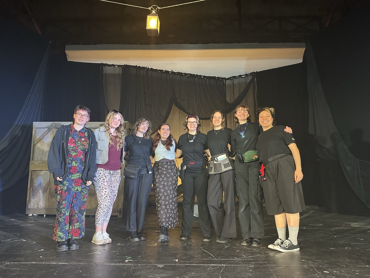 Seven people stand arm in arm on a dark stage with black curtains and a hanging light above them.