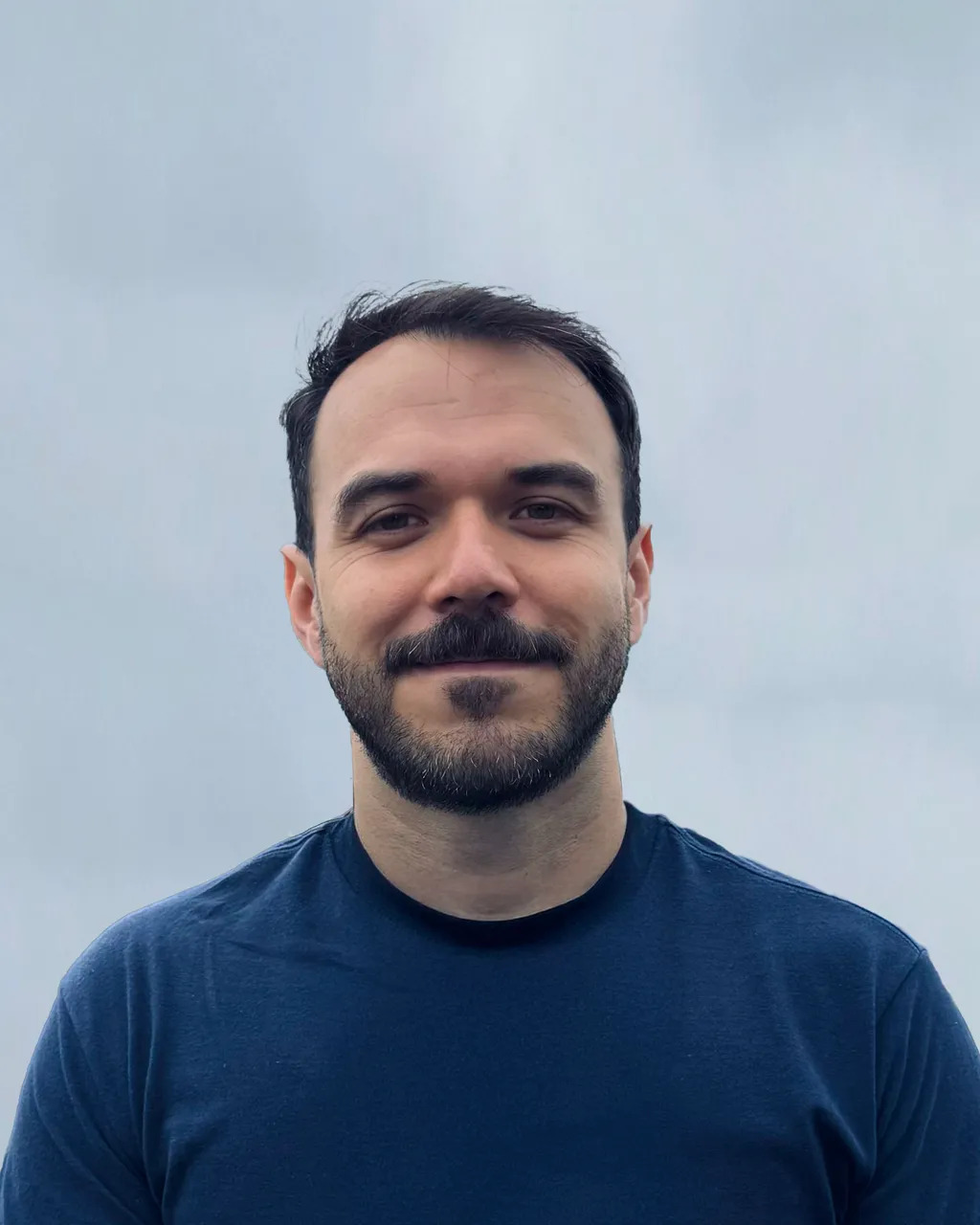 Man with dark hair and beard wearing navy blue t-shirt against light blue and purple gradient background.