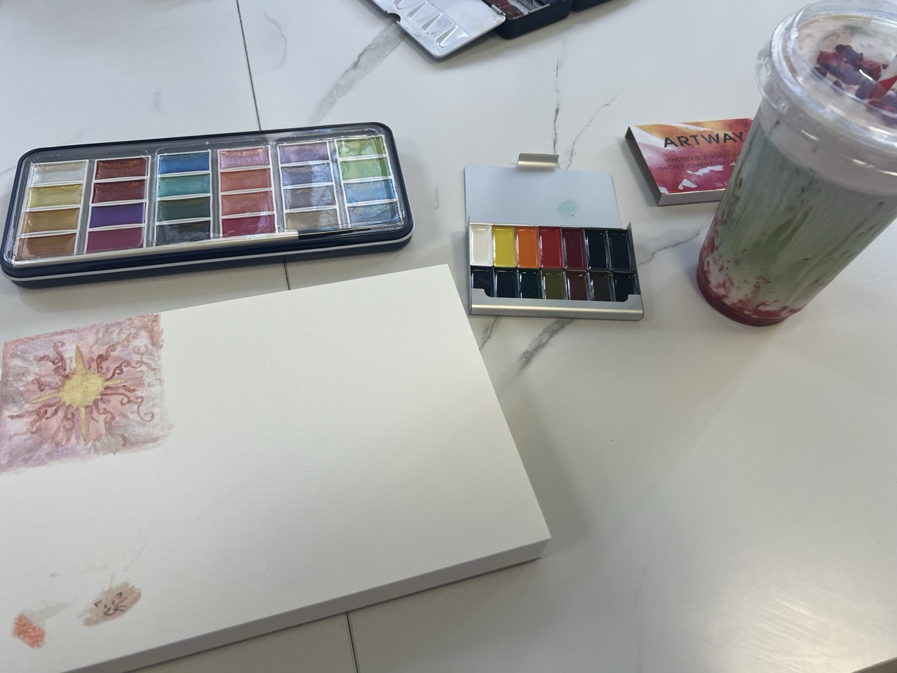 An open sketchbook with a small watercolour flower painting, beside two paint sets and a clear cup with a straw on a white table.