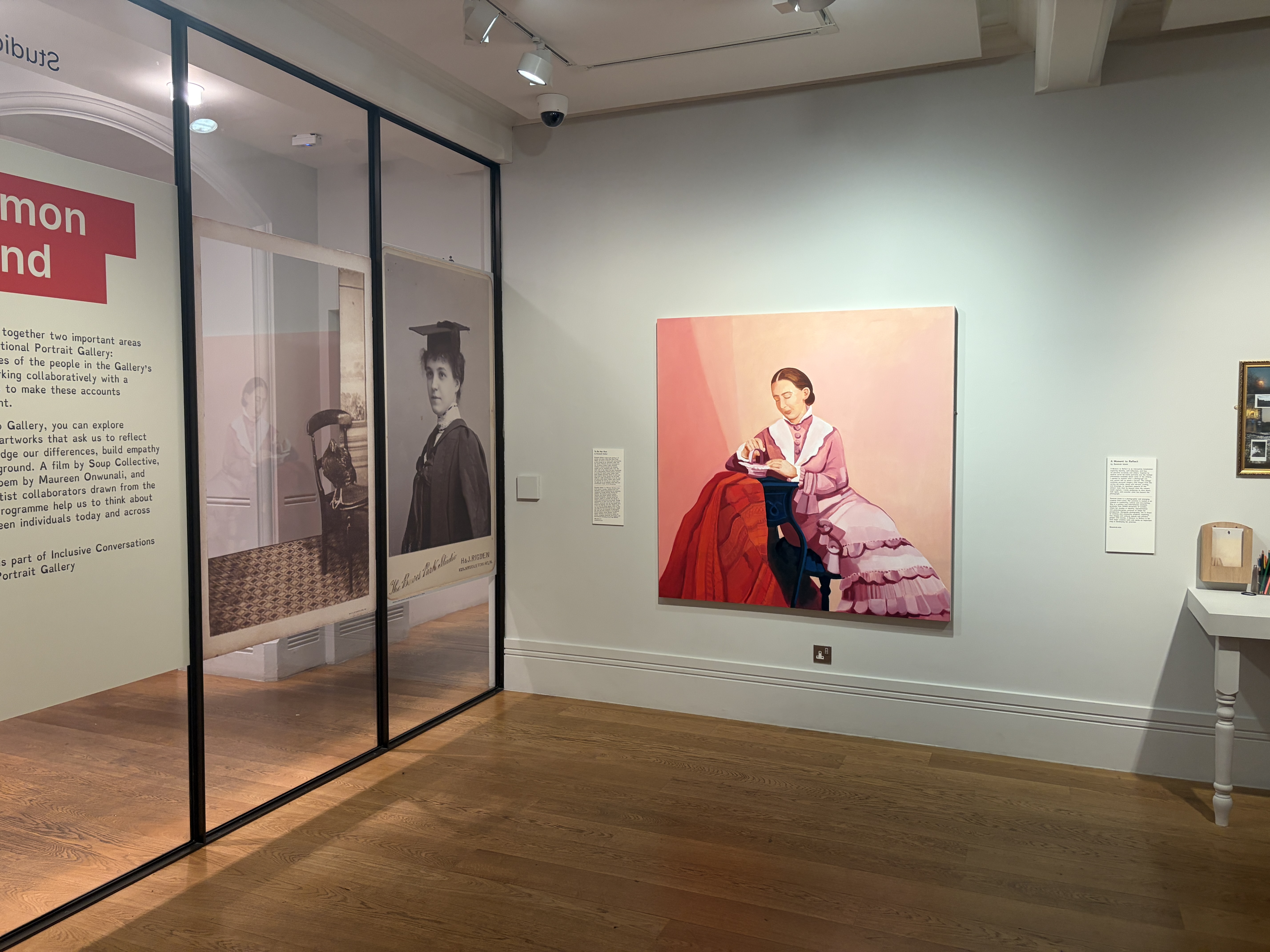 An art gallery room with a portrait of a seated woman in a pink dress on the wall, beside glass display panels and display text on a white wall.