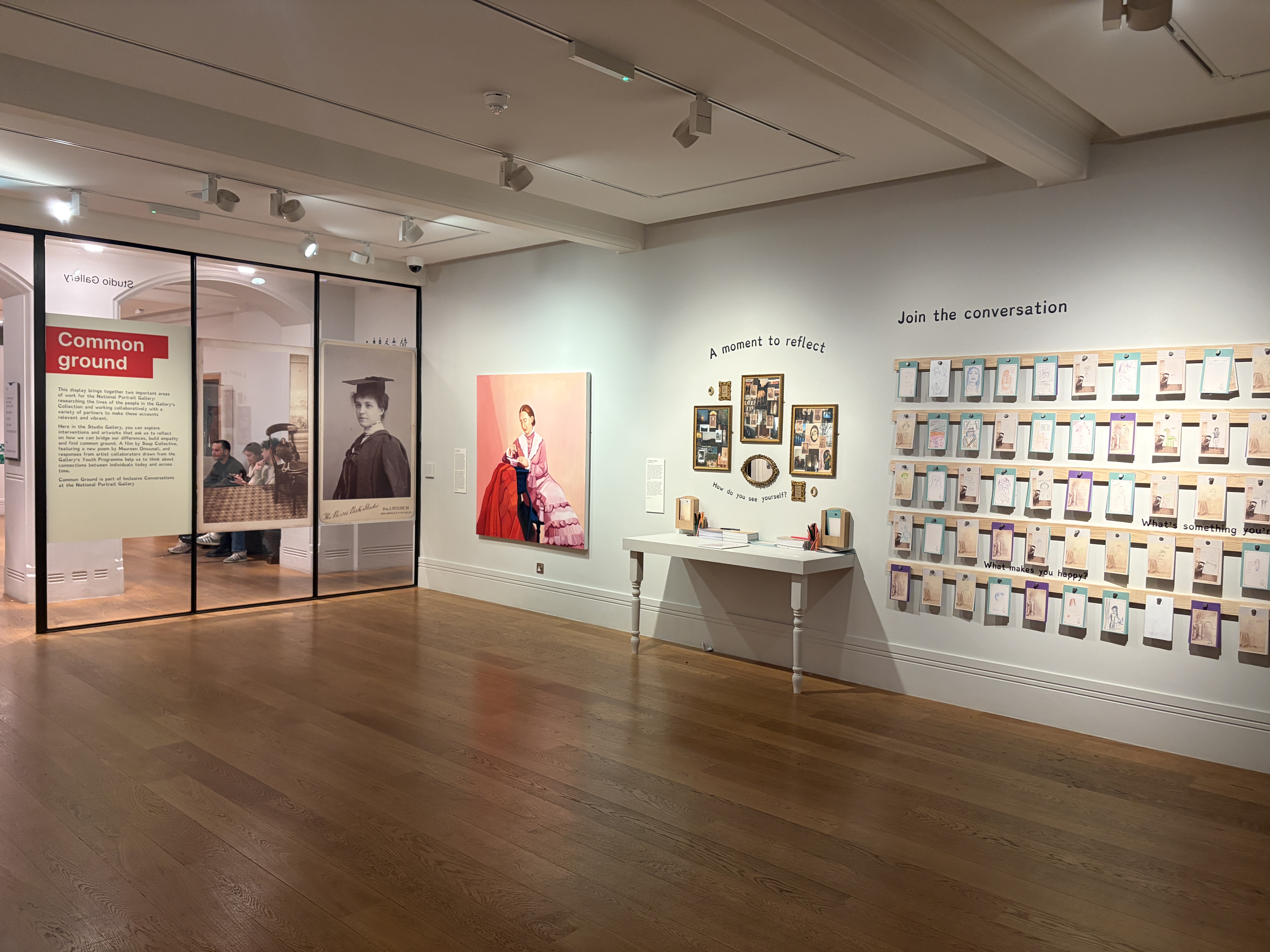 An exhibition gallery with wooden flooring, featuring framed wall displays, a table with objects and a large grid of cards under the words 'Join the conversation'.