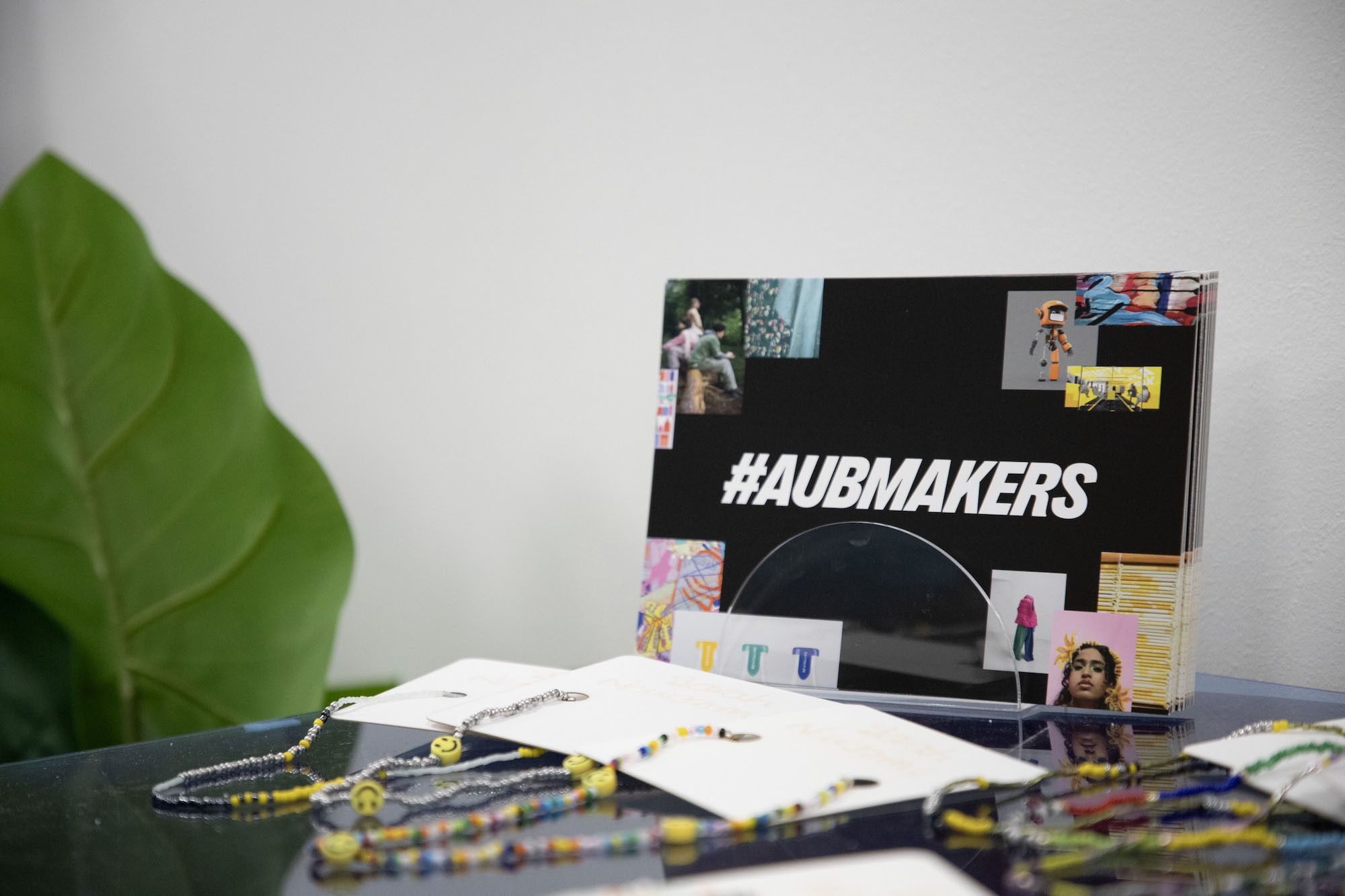 Several bead necklaces displayed on a glass table. Flyers with '#AUBMakers' written in the centre are also displayed on the table.
