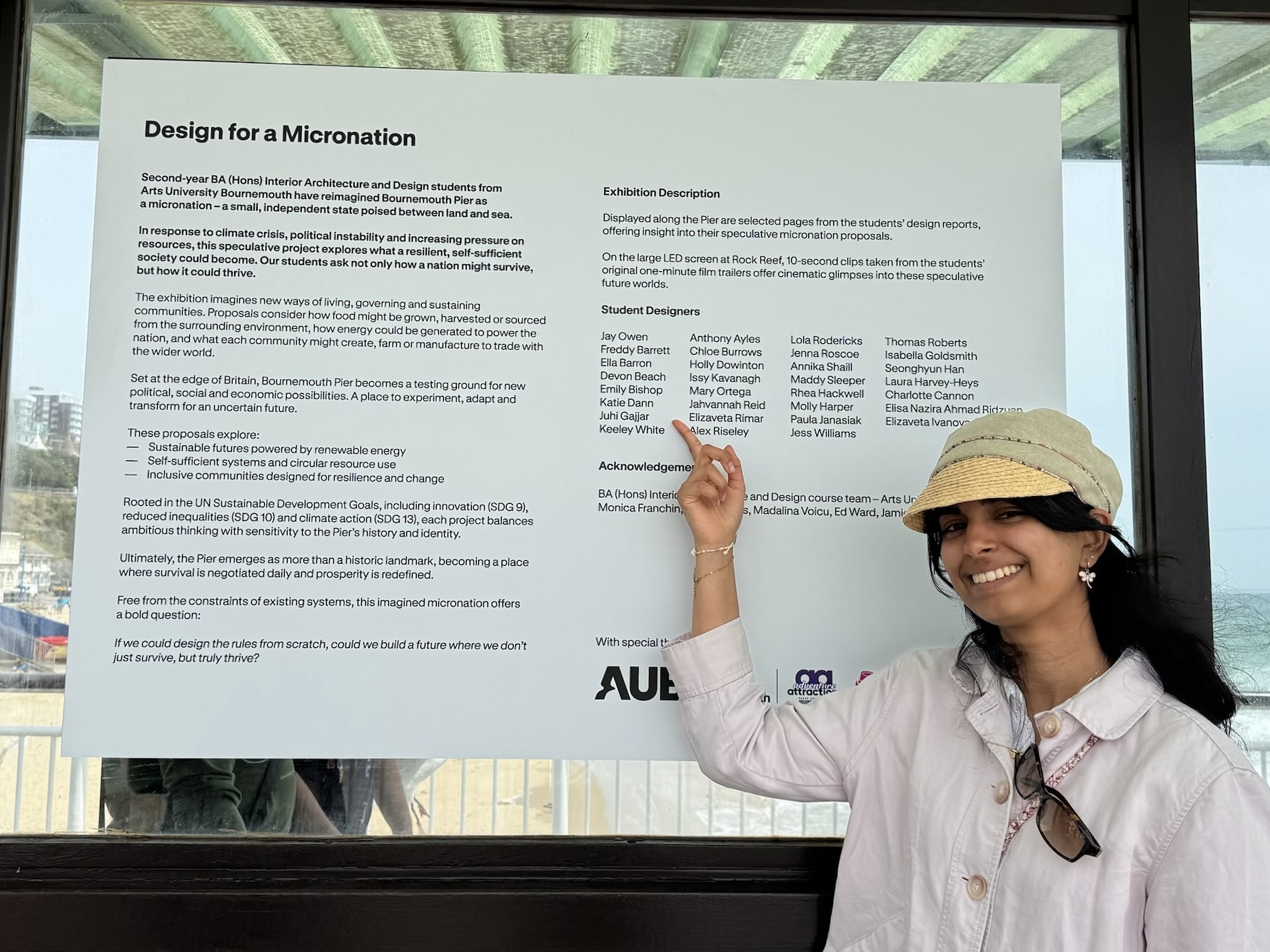 Juhi Gajjar pointing at her name on a white display talking about the project. She is wearing a hat and white shirt, smiling.