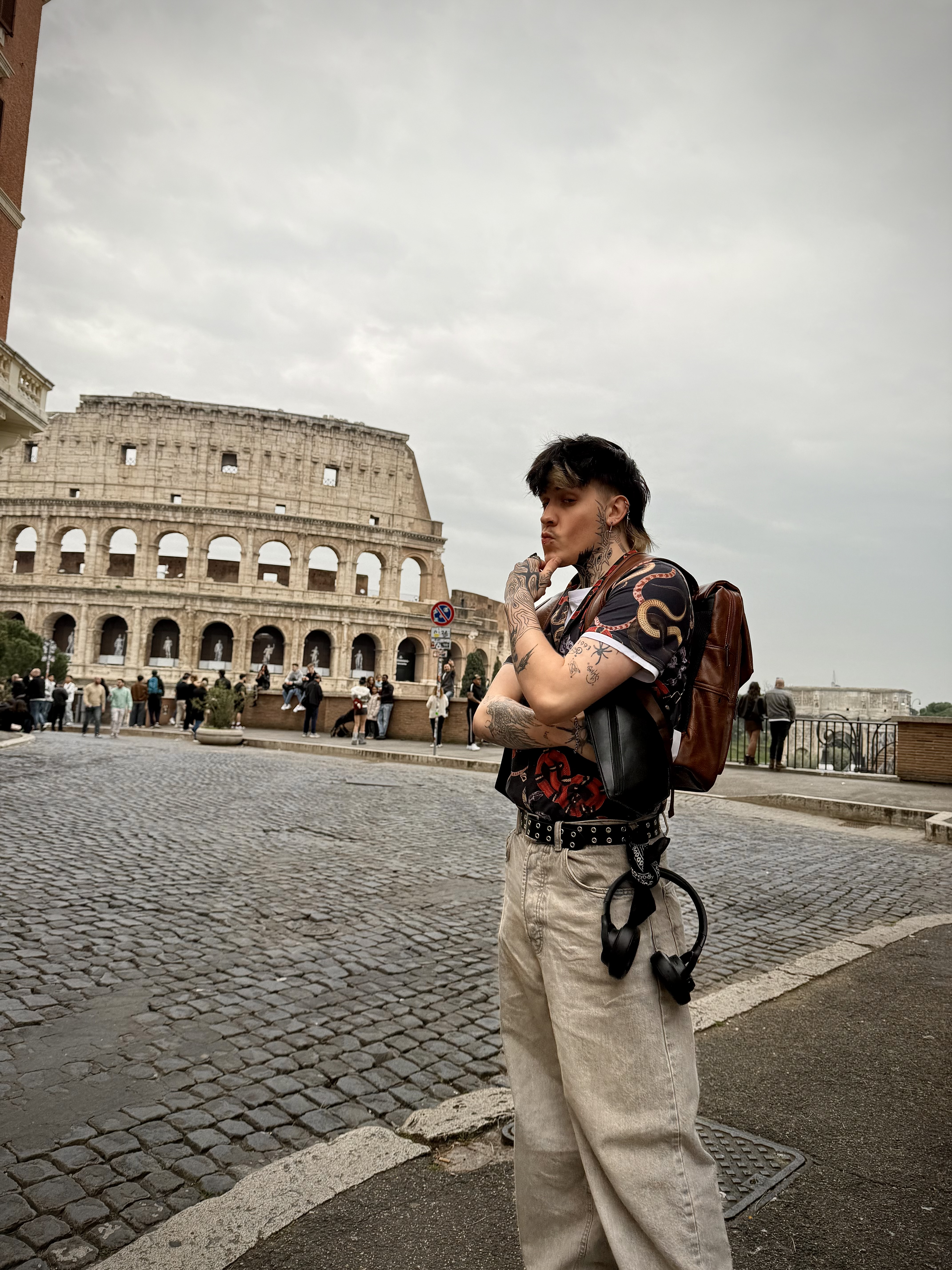 A tattooed person with a backpack and headphones stands on a cobblestone street near the Colosseum in Rome.