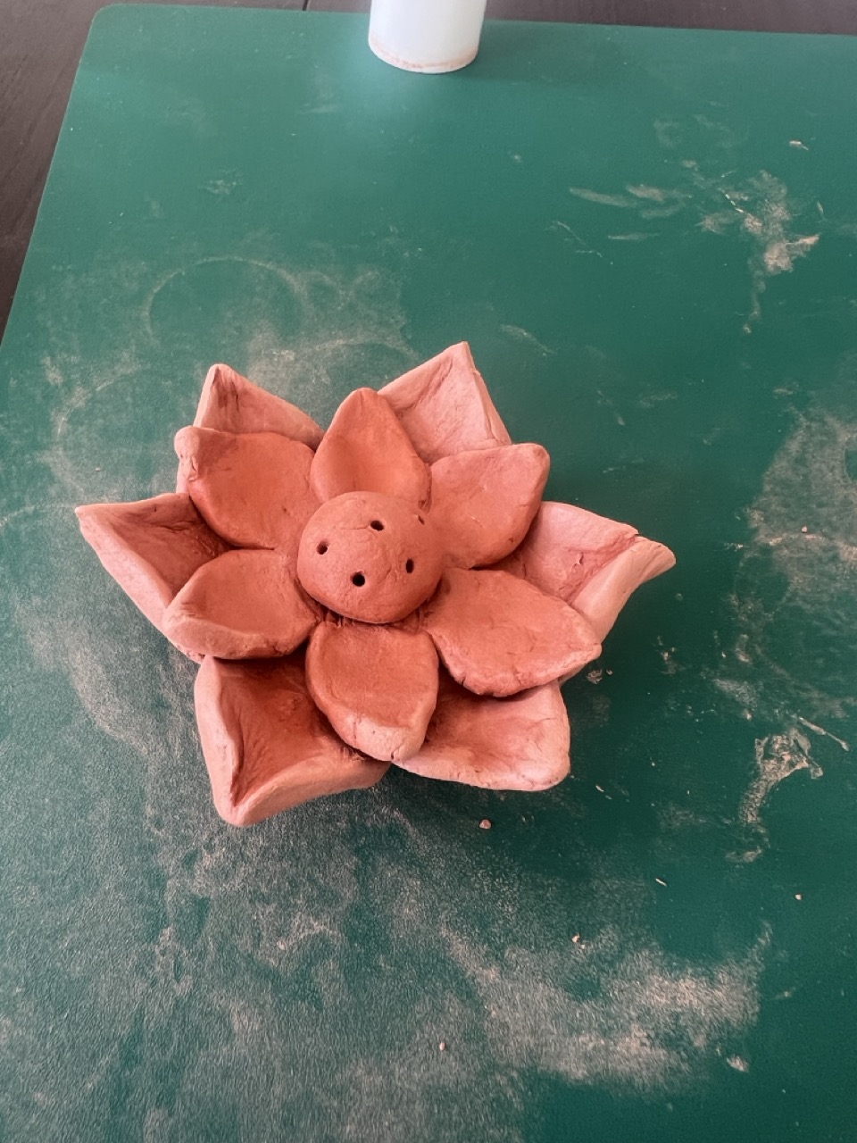 A pink clay flower sculpture with a small face sits on a green work surface, with white clay dust scattered around it.