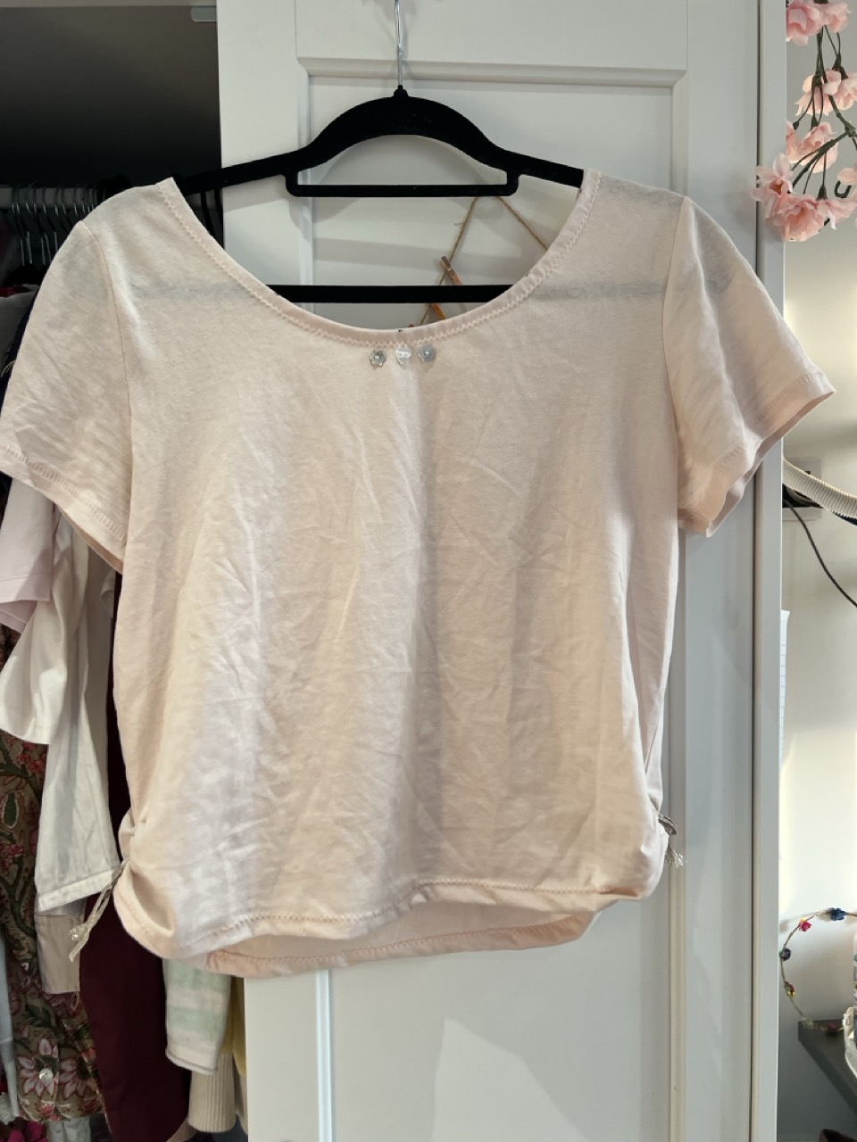 A pale pink short-sleeved T-shirt on a black hanger, hanging on a white door with other clothes partly visible behind it.