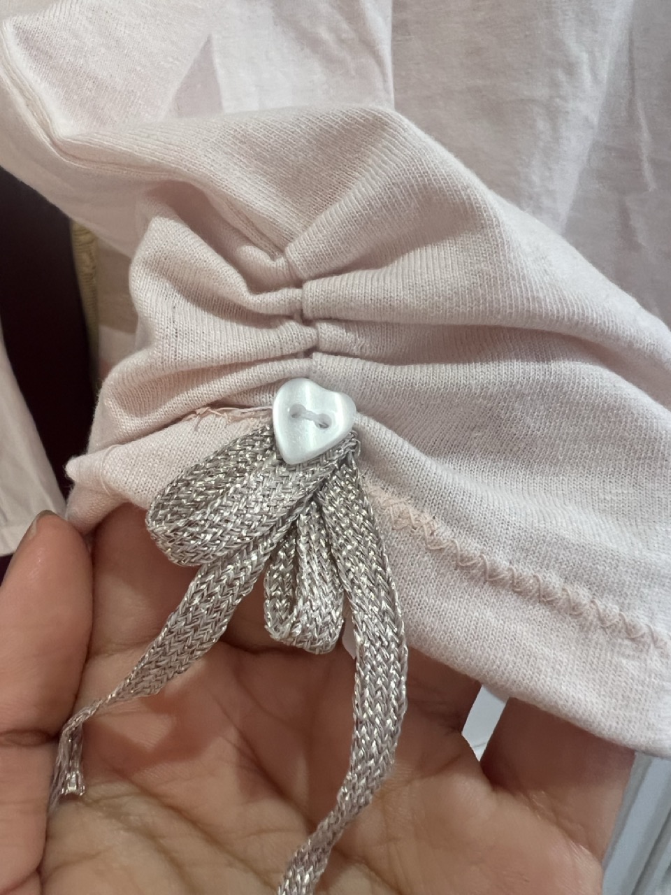 A white fabric hem with a silver woven tie and heart-shaped toggle, held by a hand, against a plain background.
