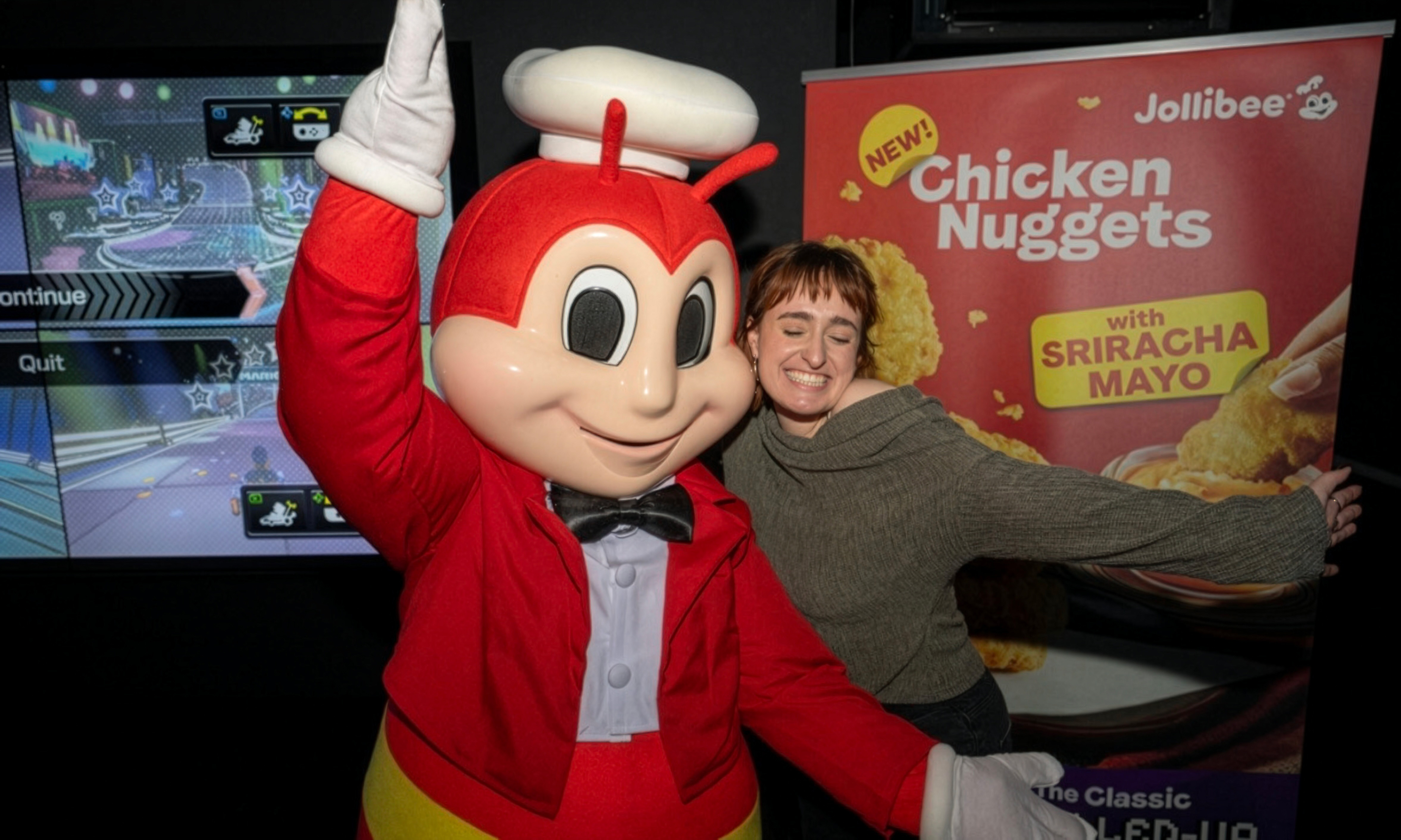 Person stood with red Jollibee mascot in a red suit, white gloves, bowtie and chef hat, Mario Kart and chicken nuggets banner behind.