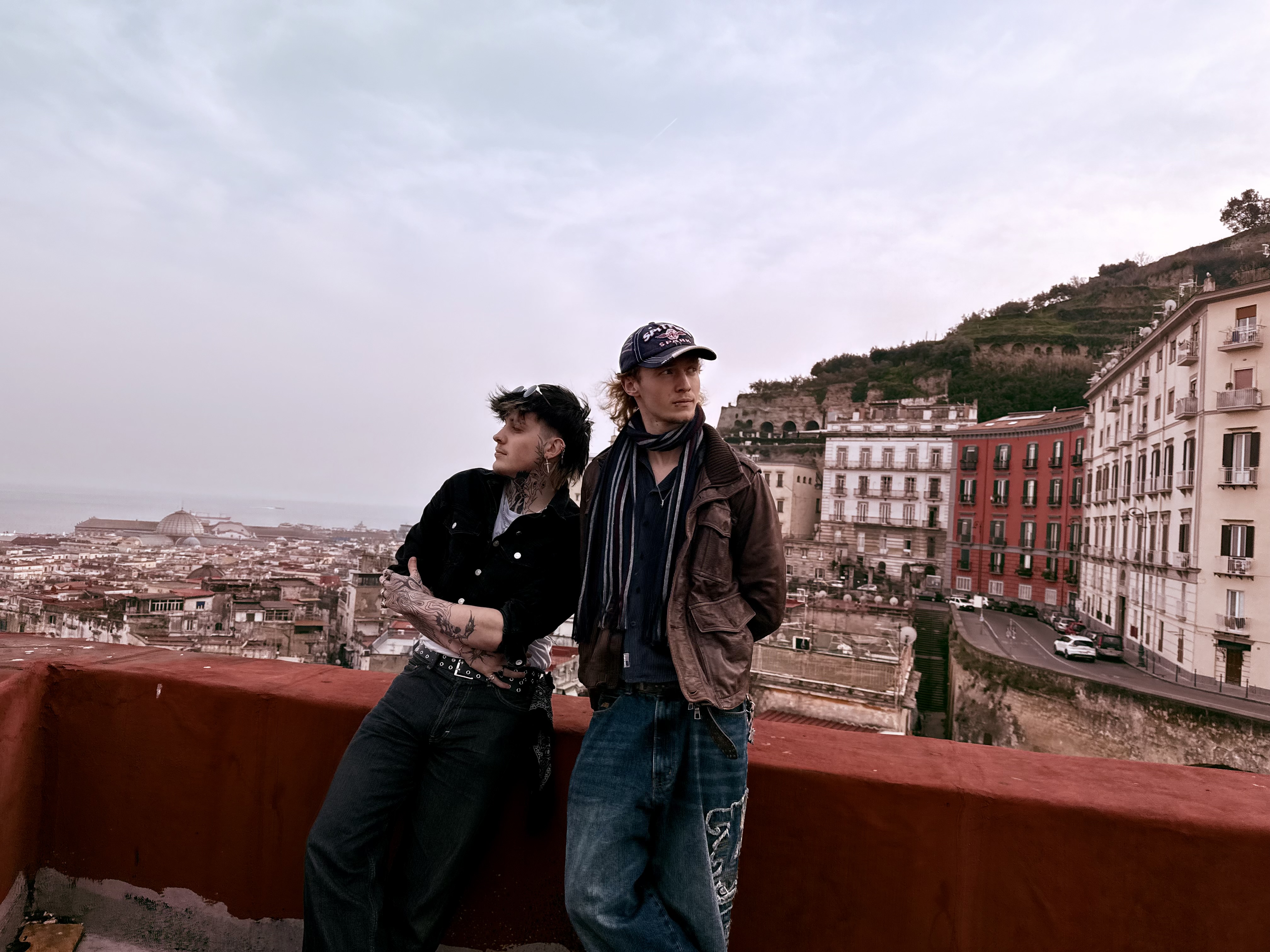 Two young men lean and stand against a red stone wall overlooking a hillside city with apartment buildings and terraced slopes.