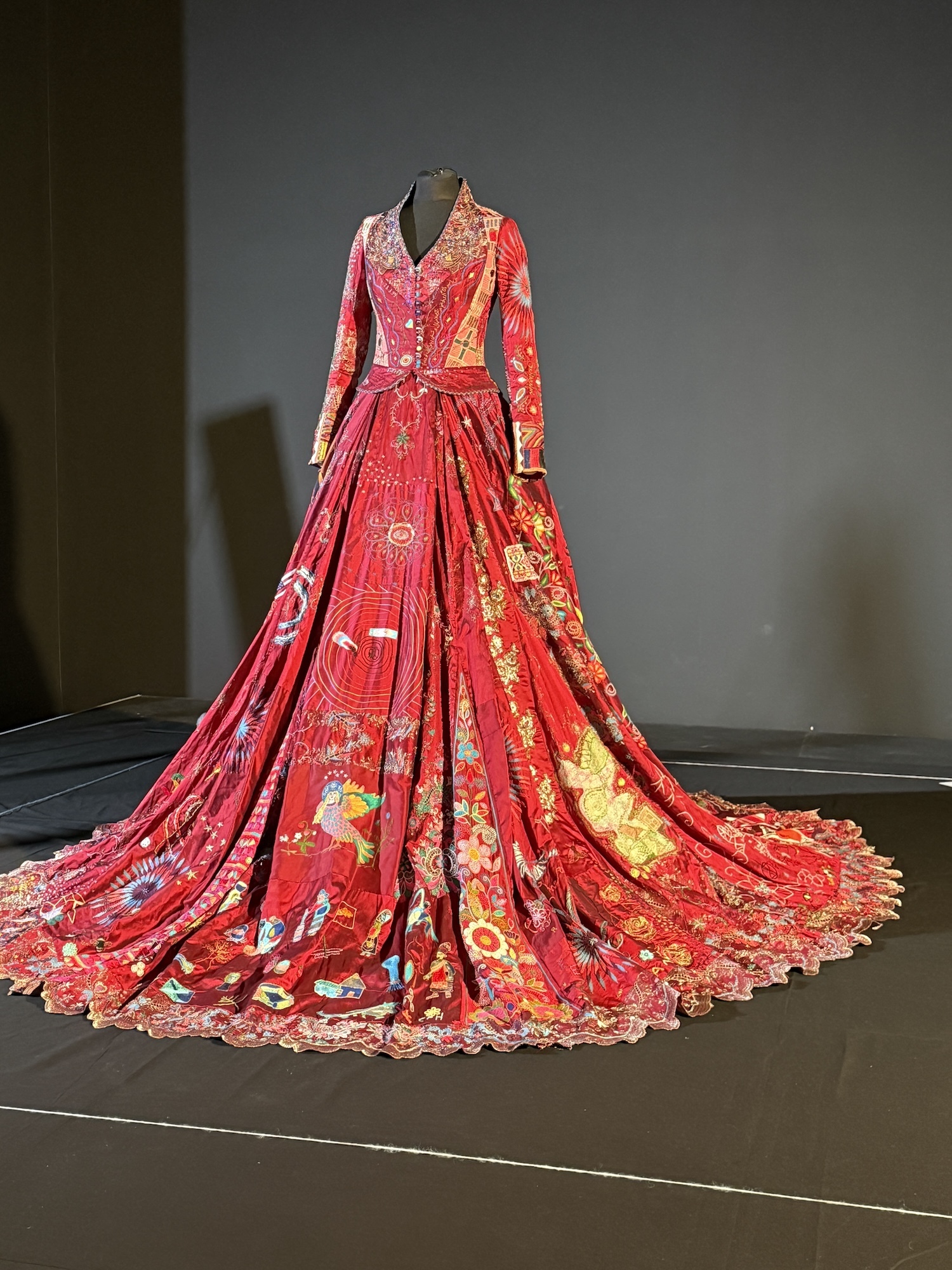 Red gown with gold embroidery and floral patterns displayed on mannequin in museum gallery with grey walls and dark flooring.