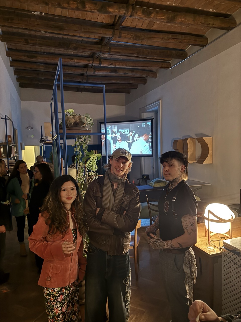 Three people standing in a warmly lit room with exposed wooden beams, a TV screen and shelving in the background.